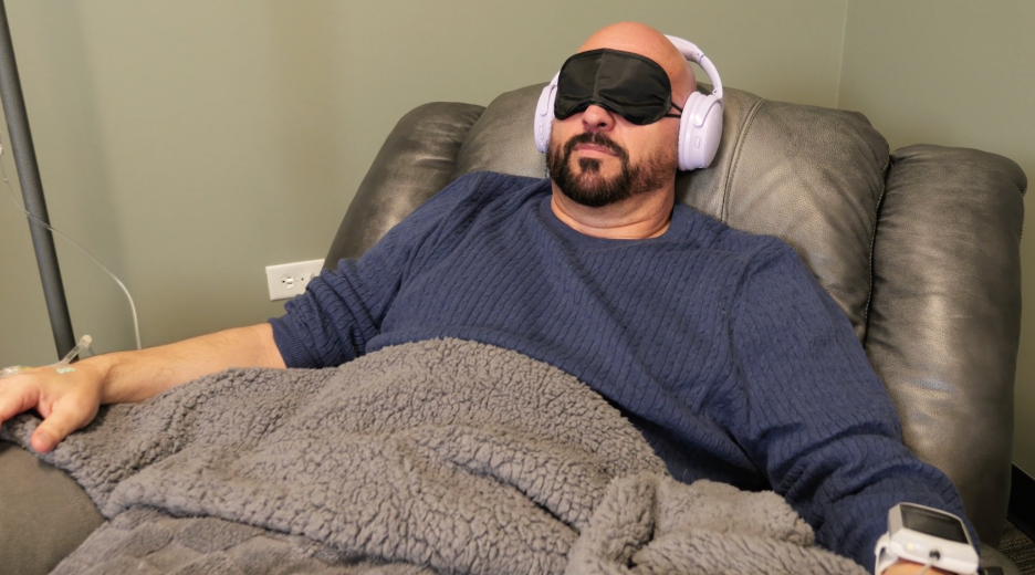 Man wearing a blindfold and headphones, resting on a recliner with a blanket, in a medical or therapy setting.