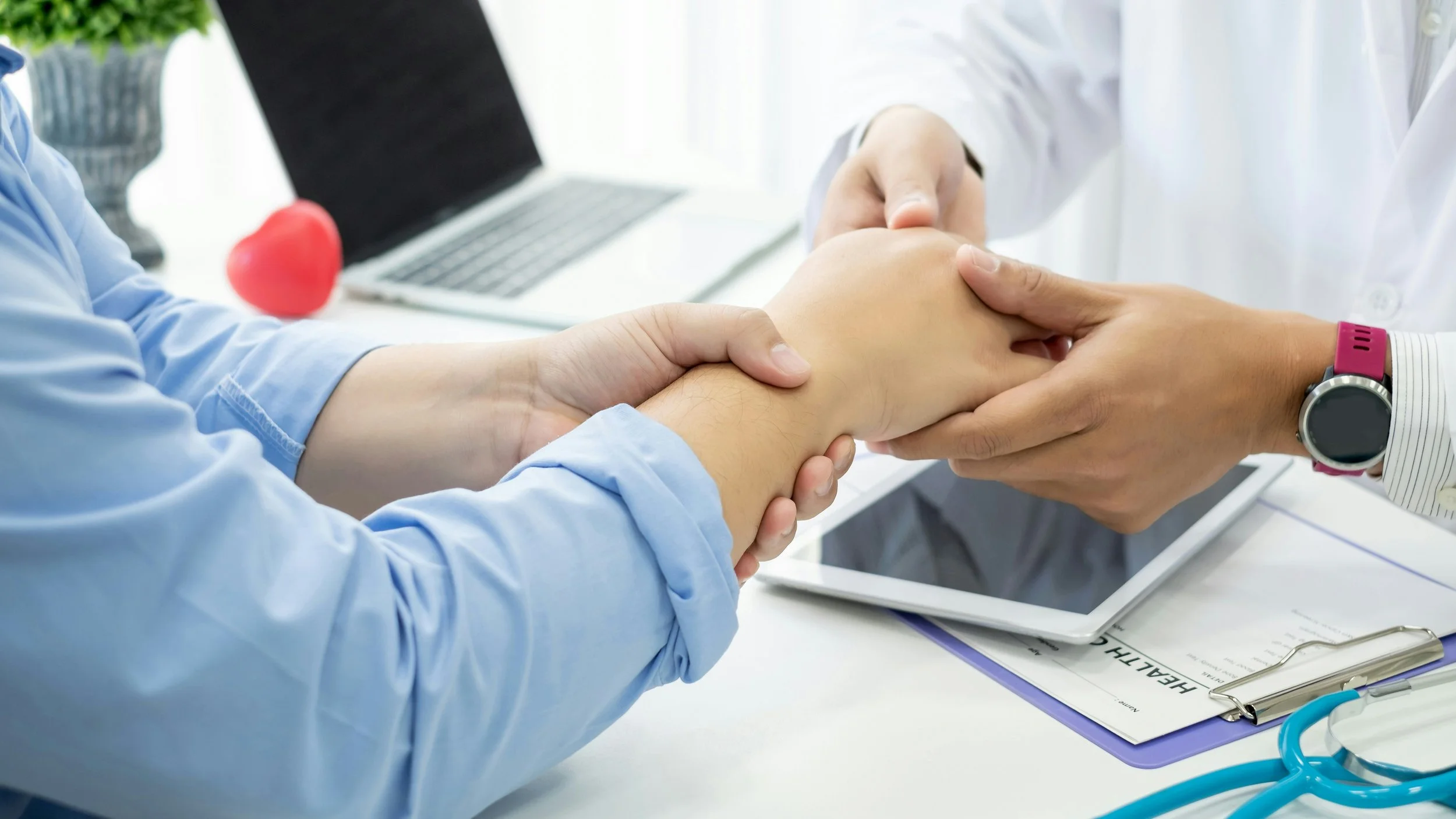 A healthcare professional in a white coat checks a patient's wrist while another person holds the patient's arm. Medical equipment, a laptop, and a clipboard are on the table.