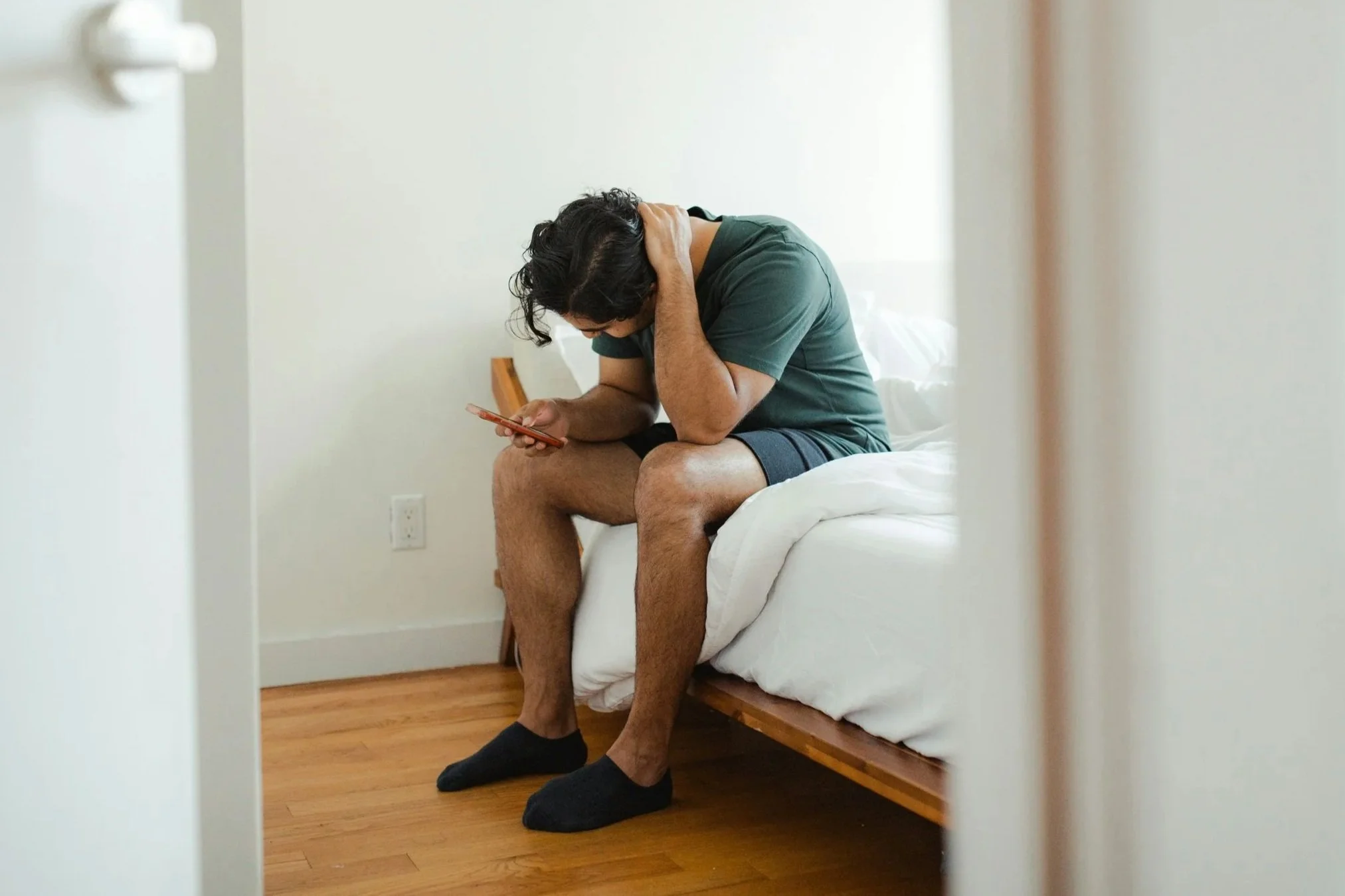 A man sits hunched on the edge of his bed, head lowered and one hand on his neck, looking down at his phone.