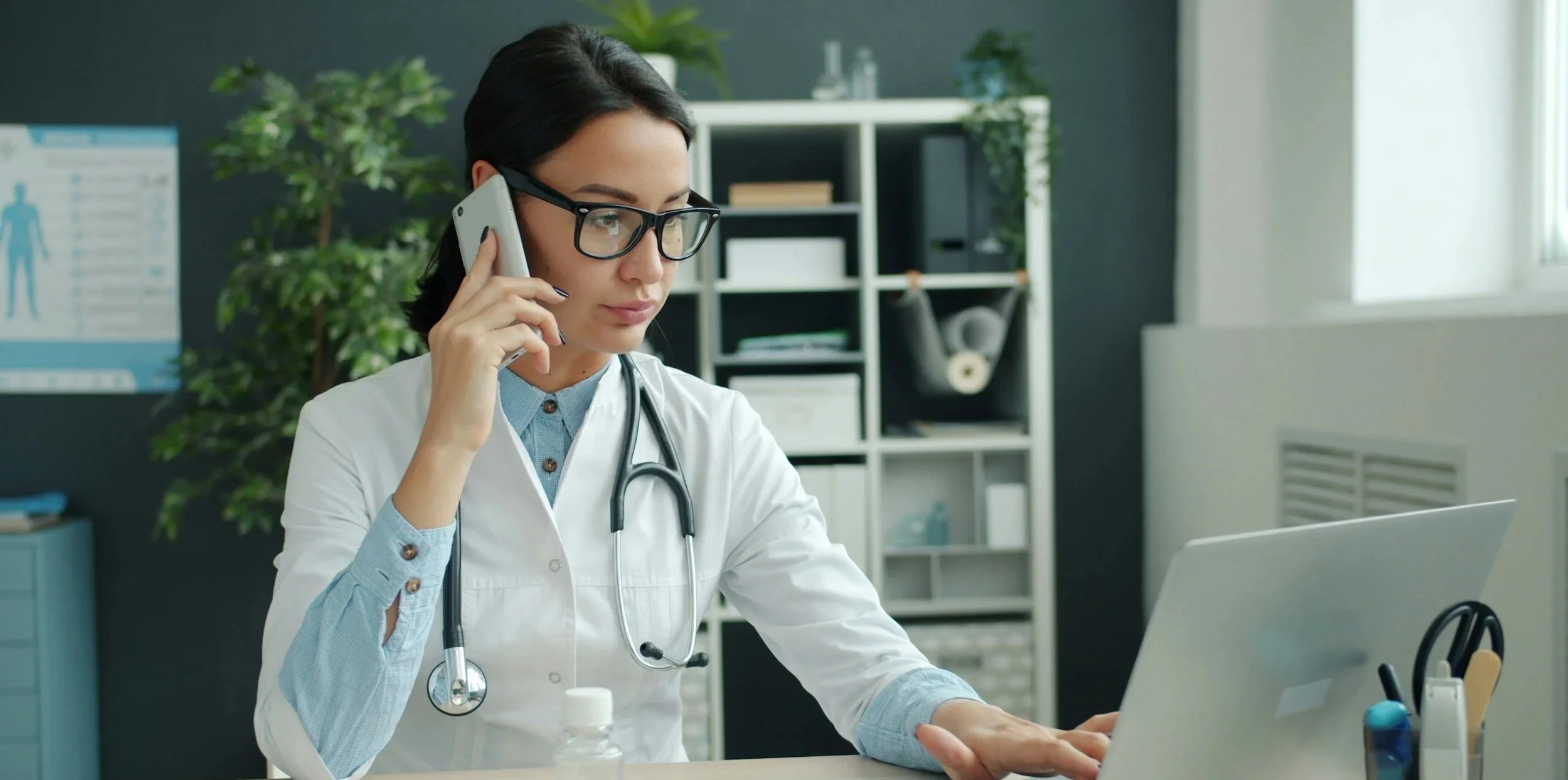 A female doctor with glasses and a stethoscope around her neck, working at a desk with a laptop, talking on a cell phone in a modern medical office.