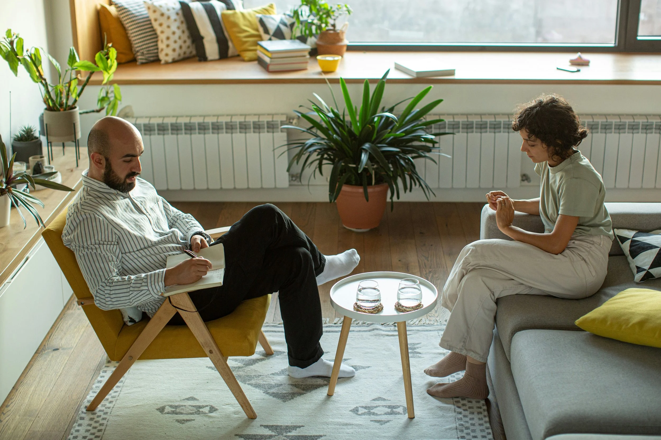 A man and a woman sitting in a living room, having a conversation. The man is seated on a yellow armchair, writing in a notebook, while the woman is seated on the sofa, listening attentively. The room is decorated with plants, pillows, and natural light coming from the large windows.
