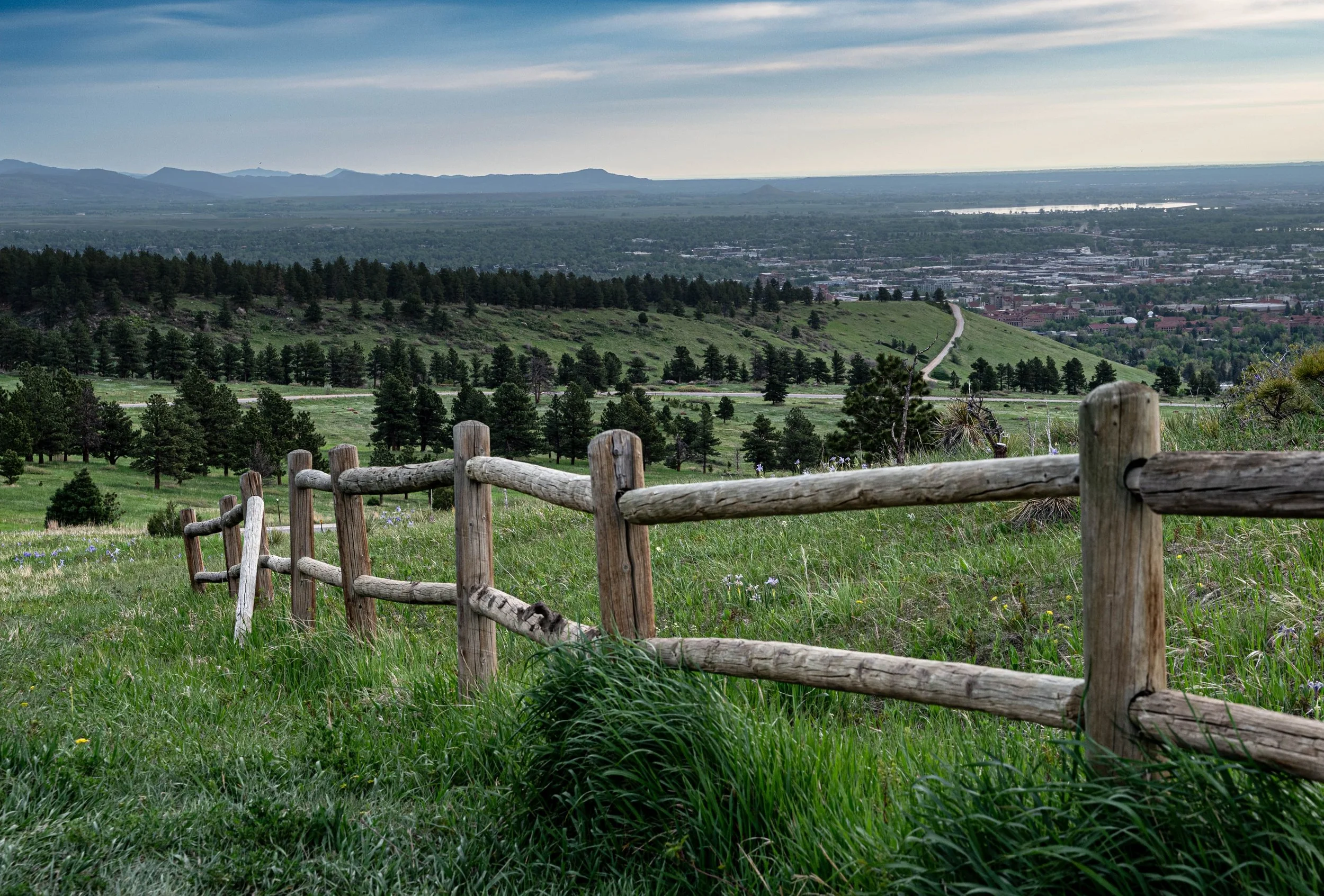 Scenic view of green rolling hills with a wooden fence, trees, and a distant city with mountains and water under a partly cloudy sky.