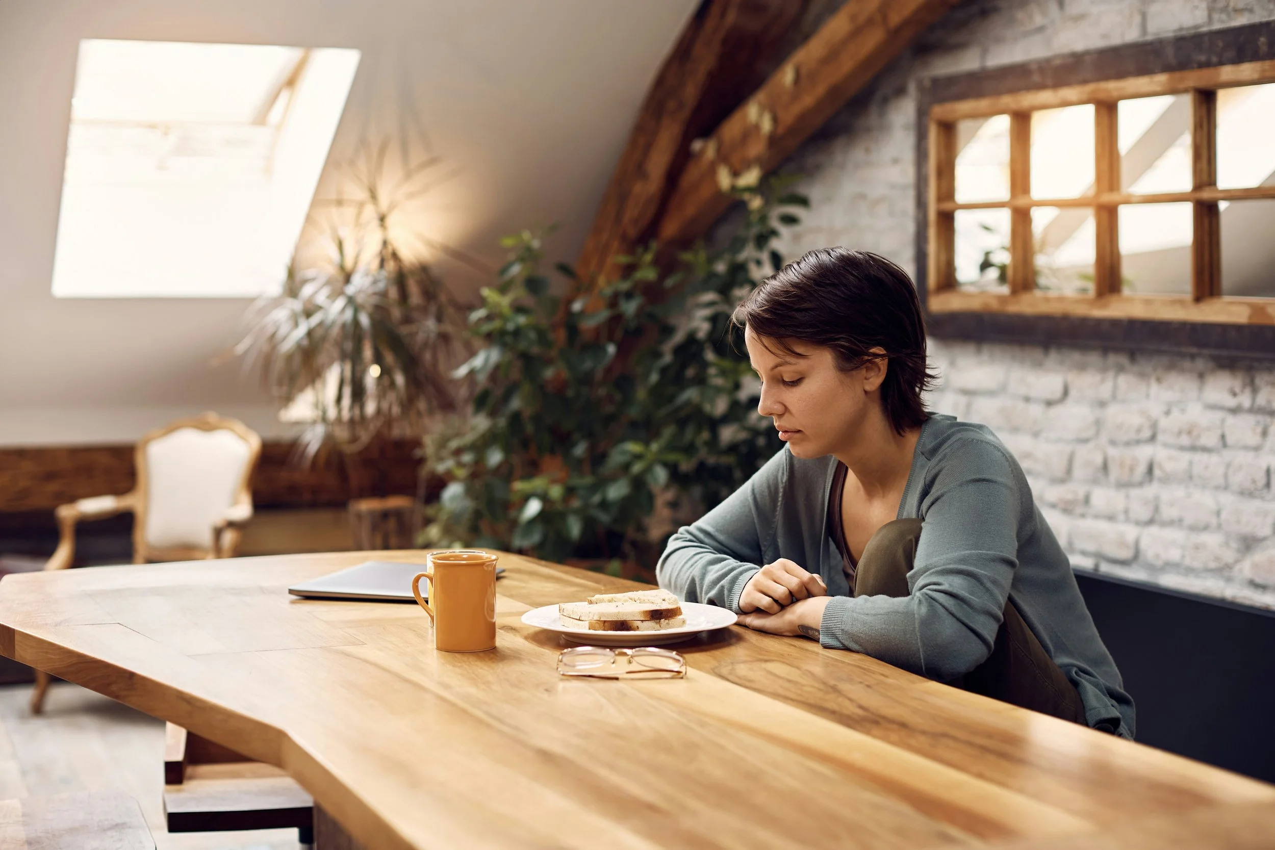 A young woman sits alone at a wooden table, looking down at an untouched plate of food, appearing quiet and withdrawn.
