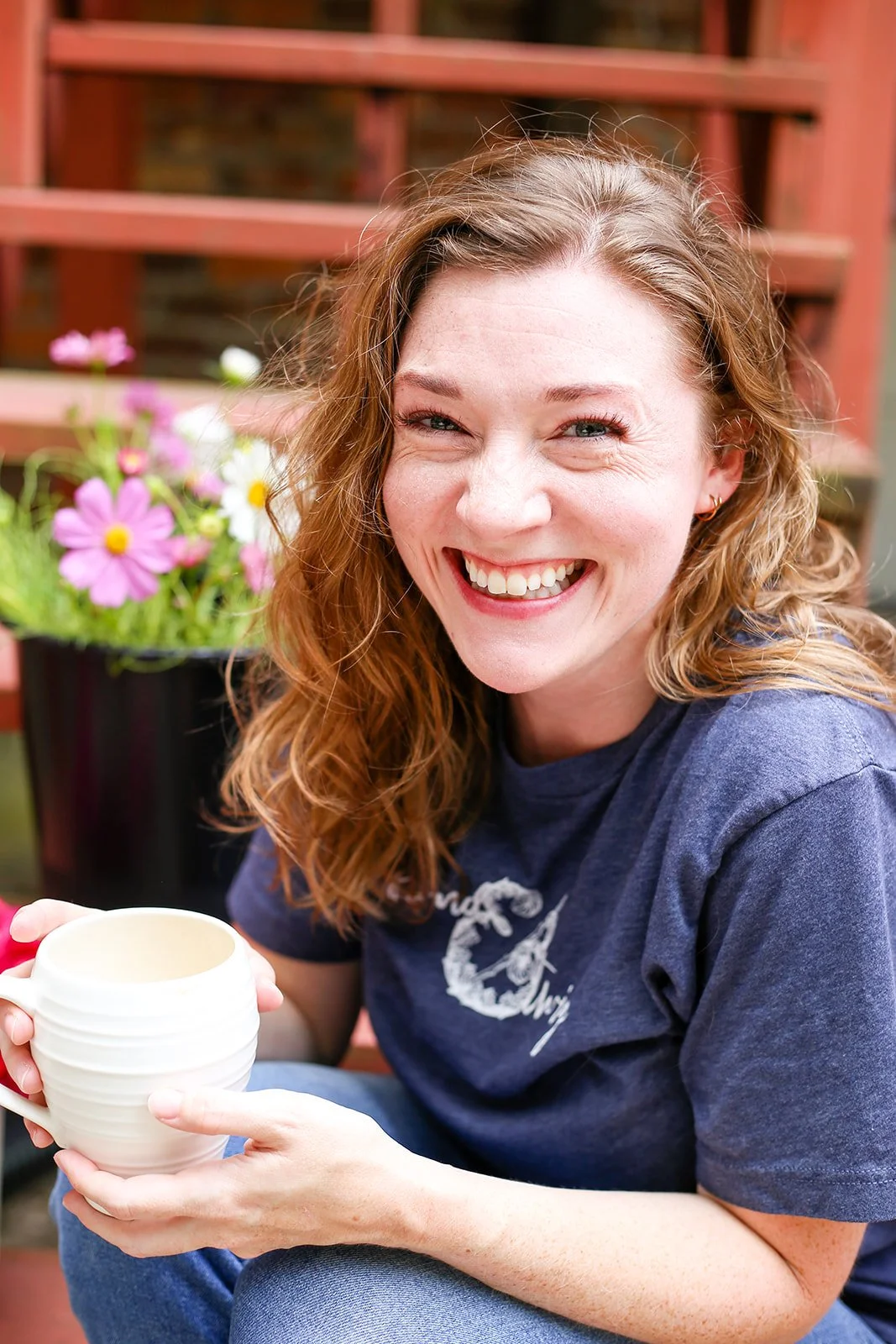 A woman with curly red hair smiling and winking, holding a mug, sitting outdoors near a flower pot with pink, white, and yellow flowers.
