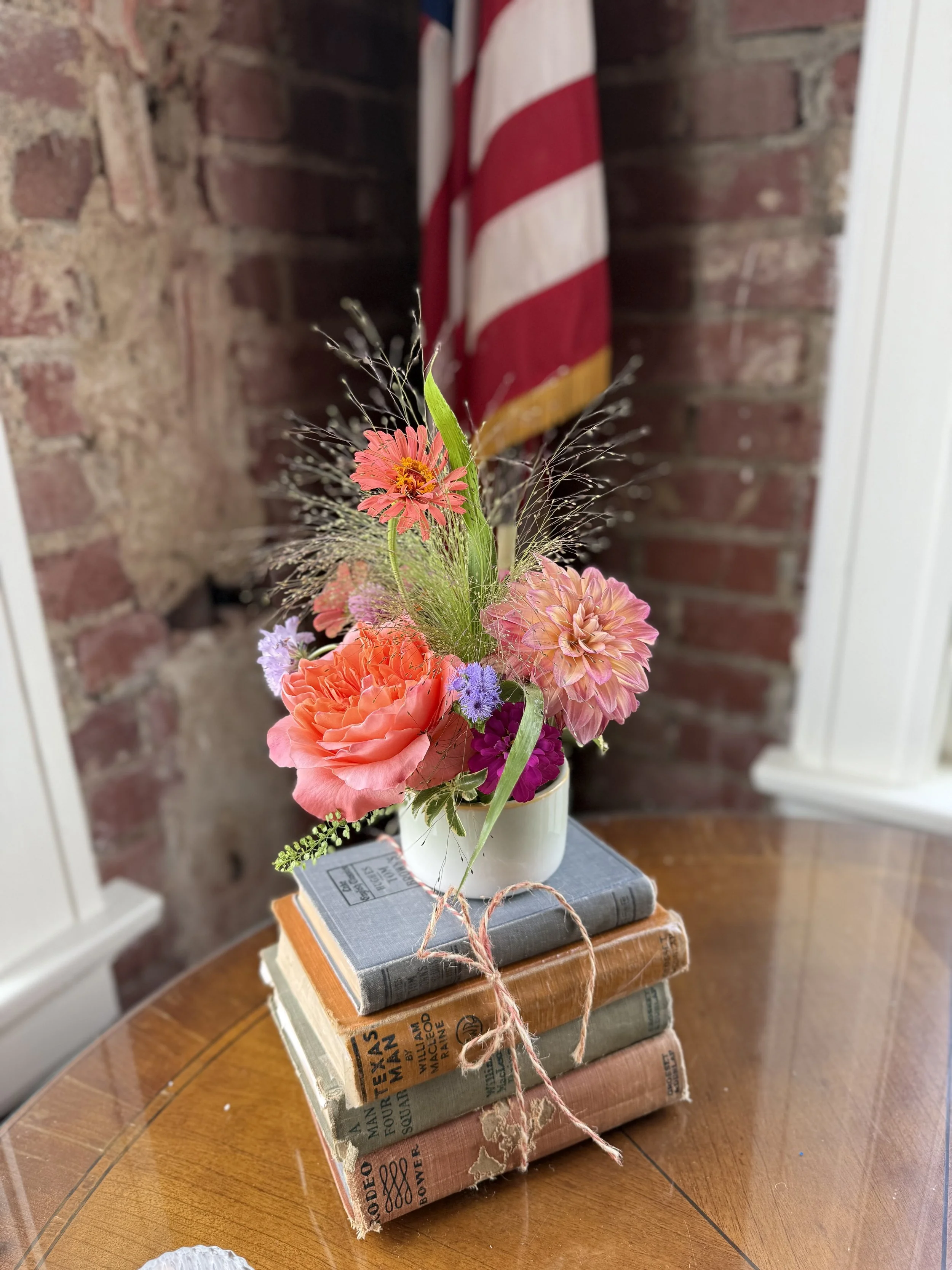 A small bouquet of pink, orange, purple, and green flowers in a white mug, placed on top of a stack of old hardcover books tied with pink string, with a brick wall and an American flag in the background.