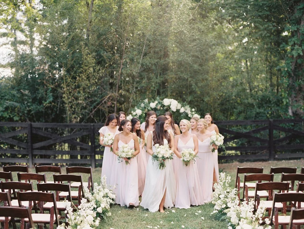 A group of women dressed in light pink and white dresses walking down an outdoor aisle decorated with white flowers, with a floral arch in the background and a black fencing on either side of the aisle.