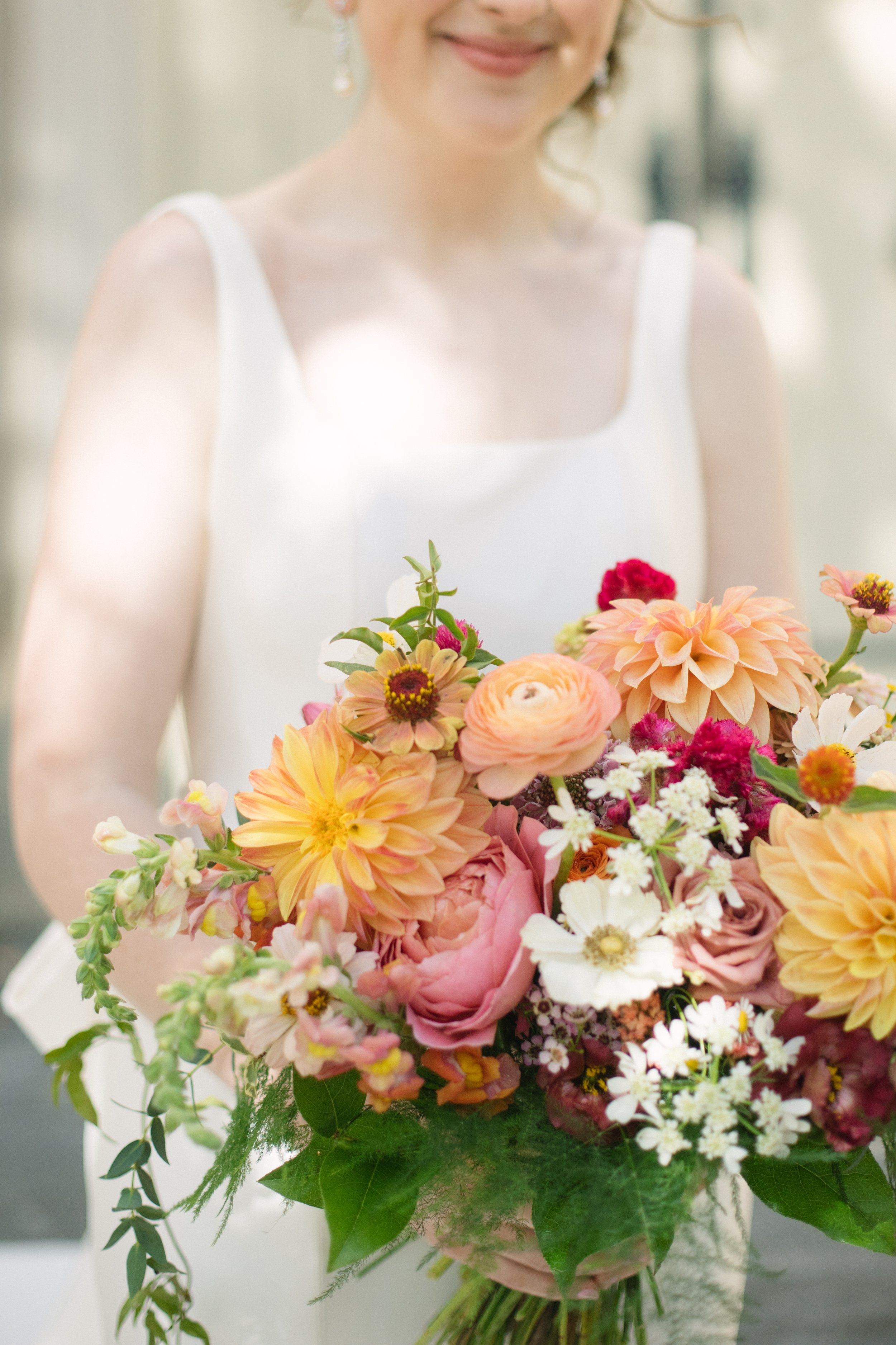A woman in a white sleeveless dress holding a large bouquet of pink, peach, and white flowers with green leaves.