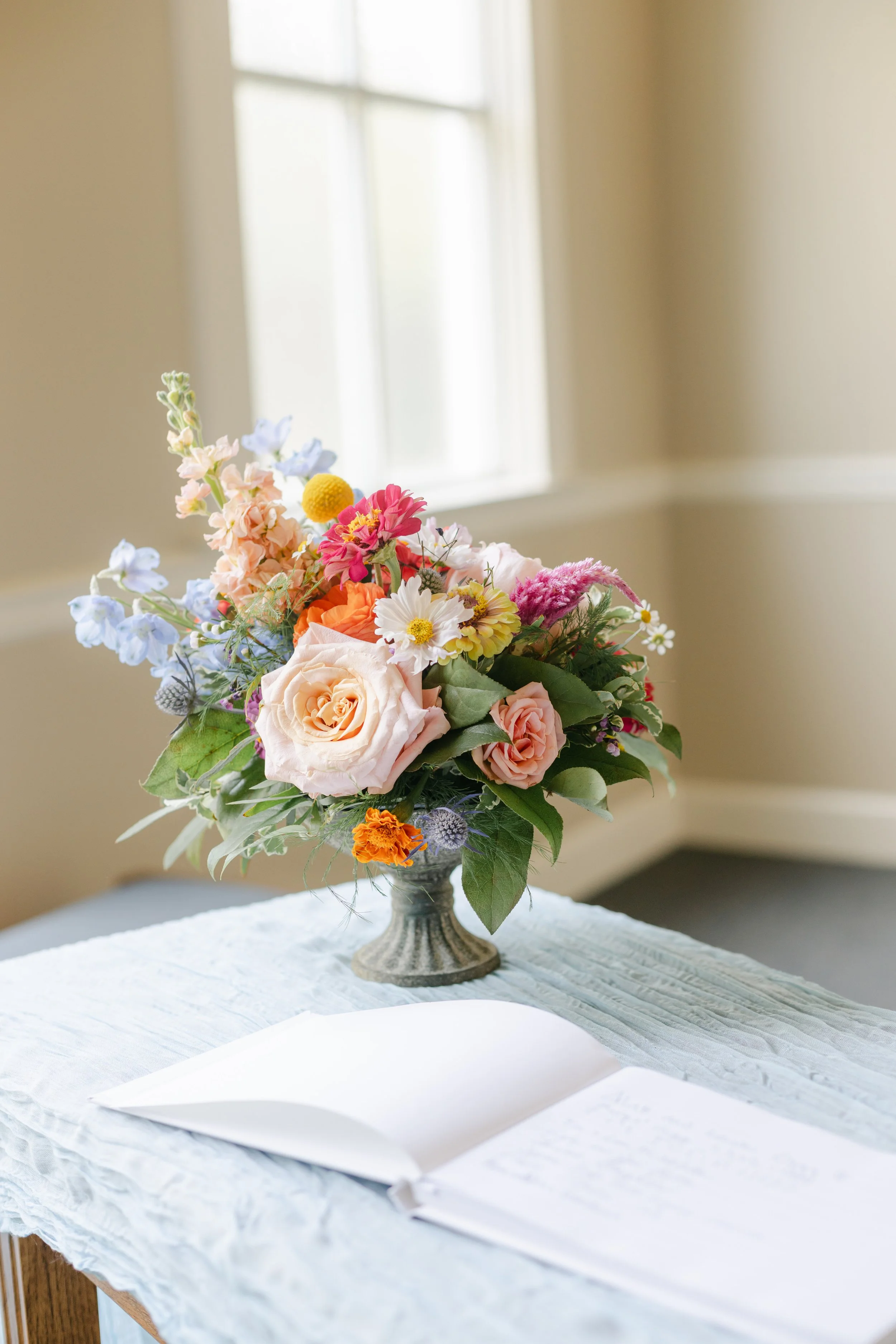 A colorful flower bouquet in a small pedestal vase, placed on a light-colored table with a white cloth, and an open guestbook with handwritten notes in front of the flowers. A window with natural light is seen in the background.