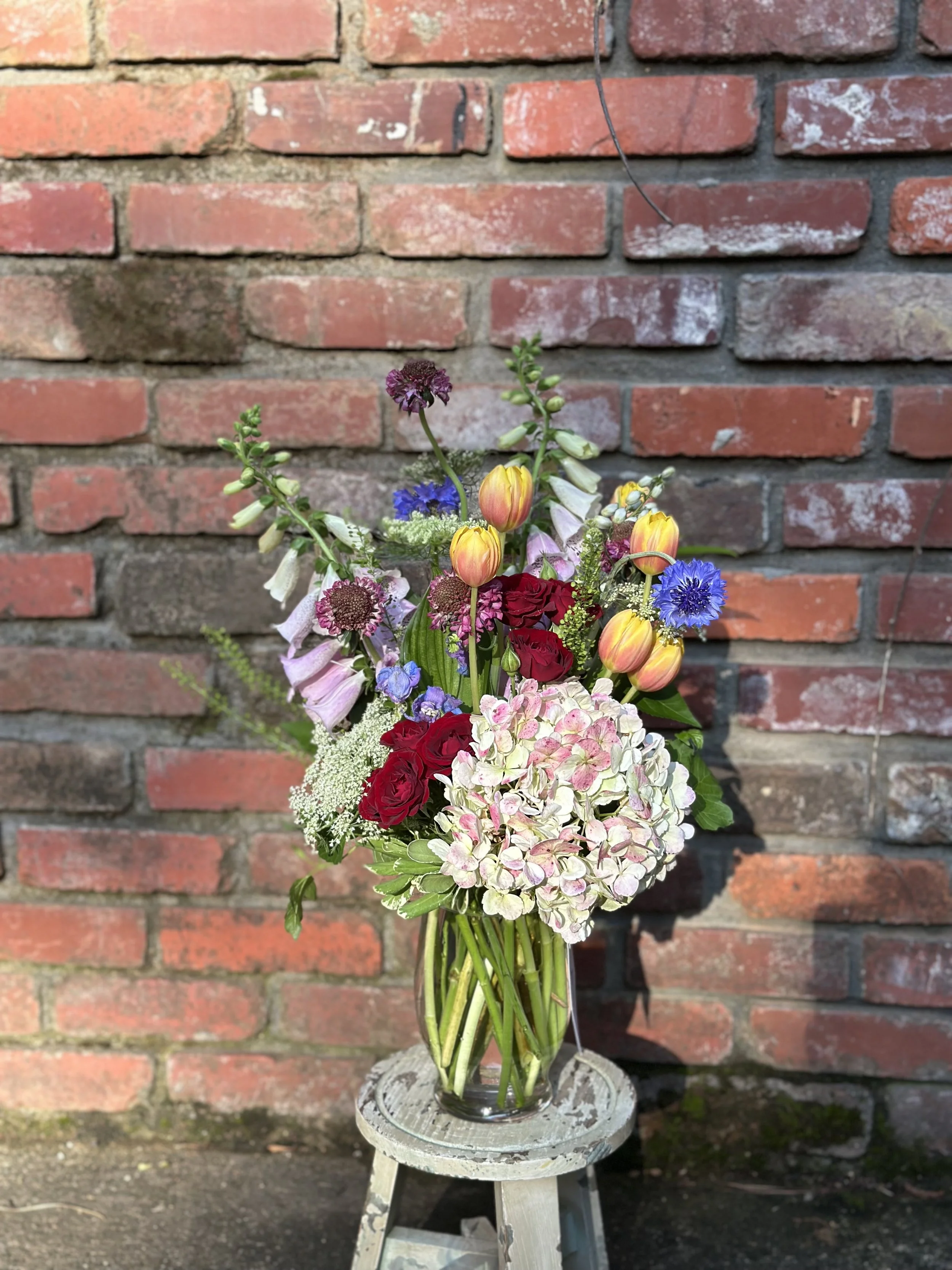 A colorful bouquet of flowers in a glass vase on a small white stool against a brick wall background.