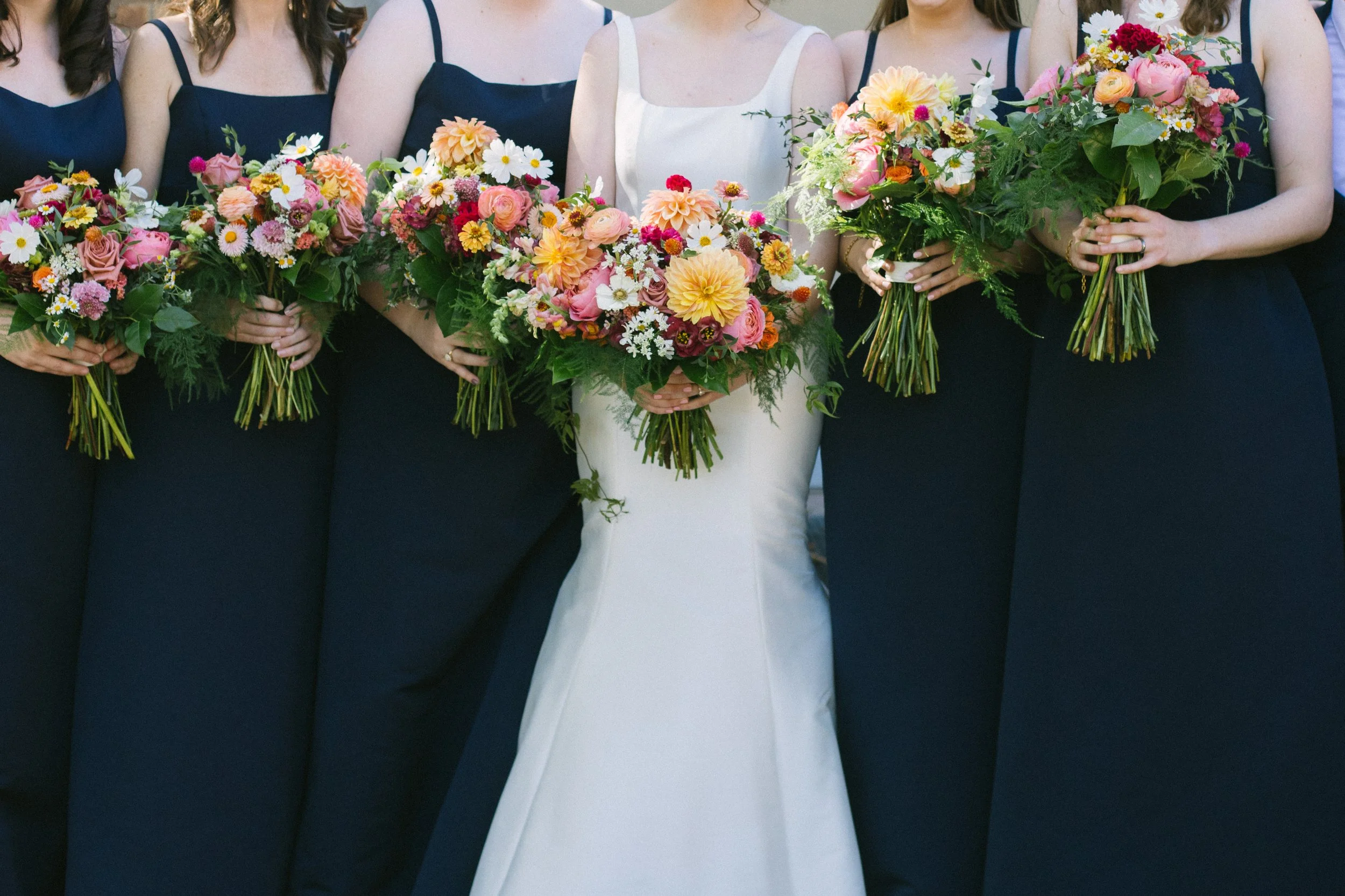 A bride in a white wedding dress holding a large bouquet of colorful flowers, standing among bridesmaids dressed in navy blue gowns, each holding a bouquet of mixed flowers.
