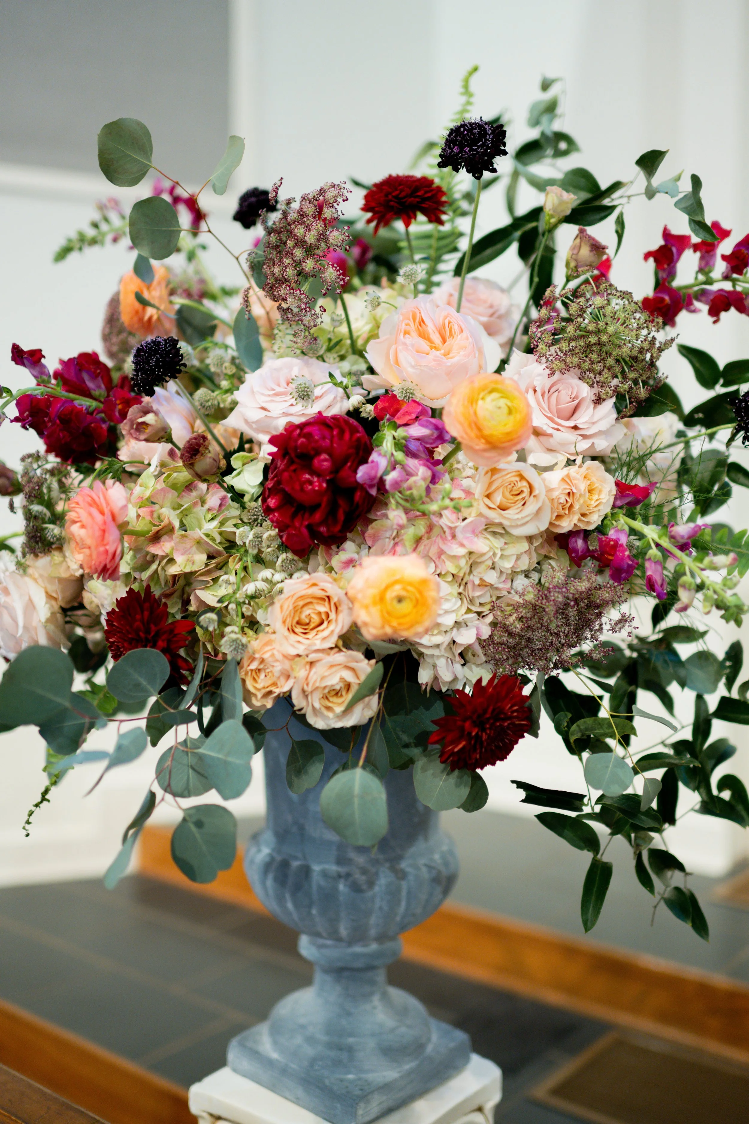 Elegant flower arrangement with roses, ranunculus, dahlias, and greenery in a classical urn vase on a wooden table.