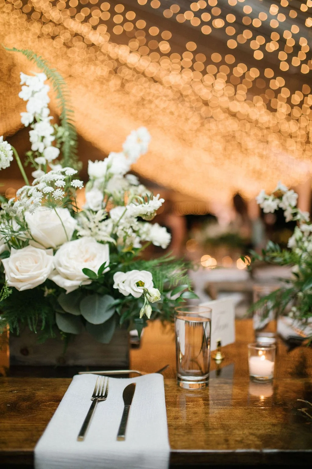 A table setting at a wedding reception with a floral centerpiece of white roses and greenery, a glass of water, a lit candle, and a white napkin with fork and knife, with warm blurred string lights in the background.