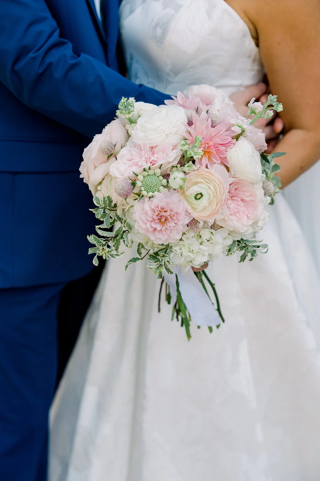 Close-up of a wedding bouquet with pink and white flowers held by a bride, while a groom in a blue suit stands beside her.