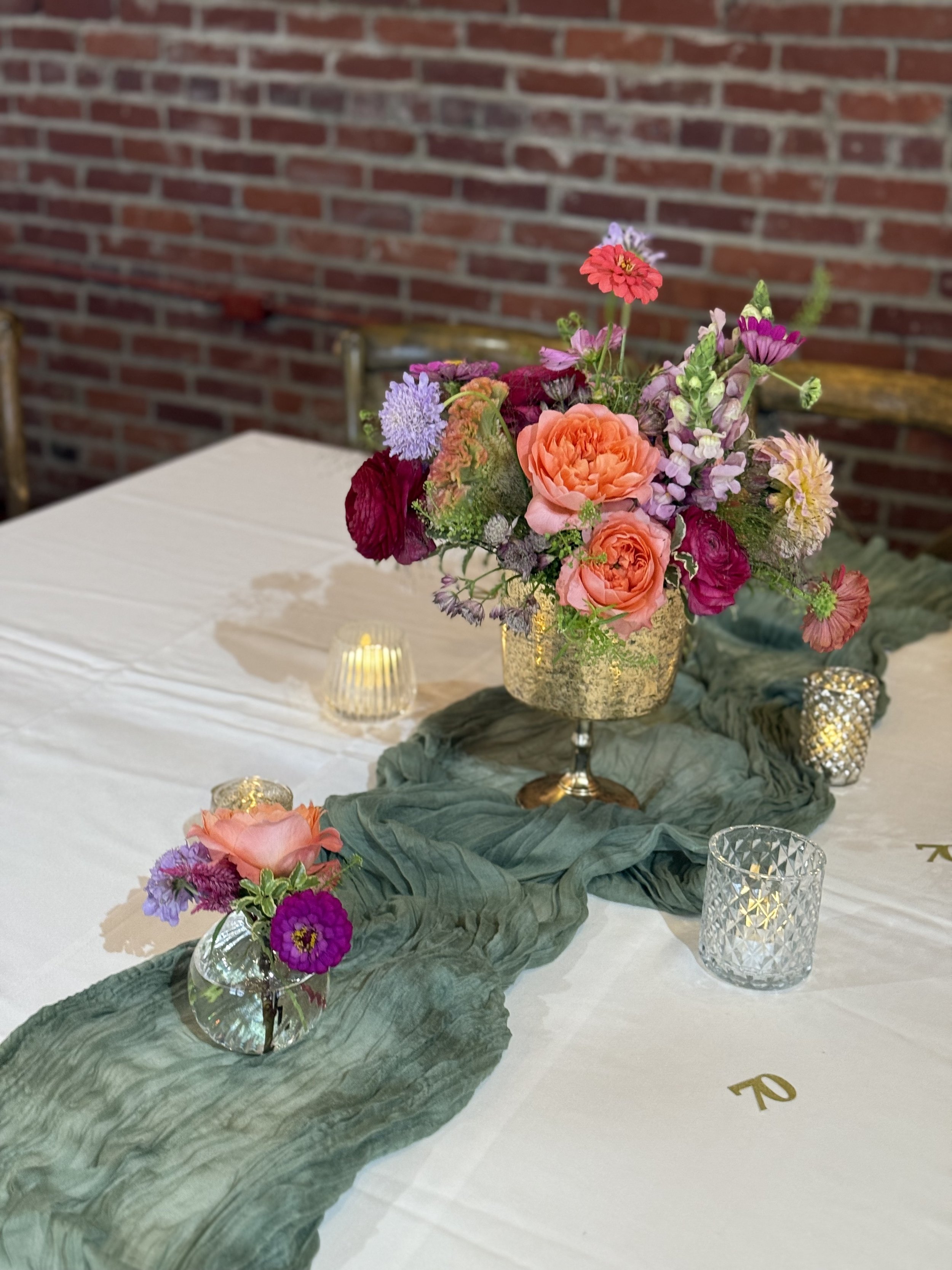 A table decorated with a green table runner, a large gold vase with pink, purple, and red flowers, smaller glass votive candle holders, and a small glass vase with a single pink flower and purple accents, against a brick wall background.