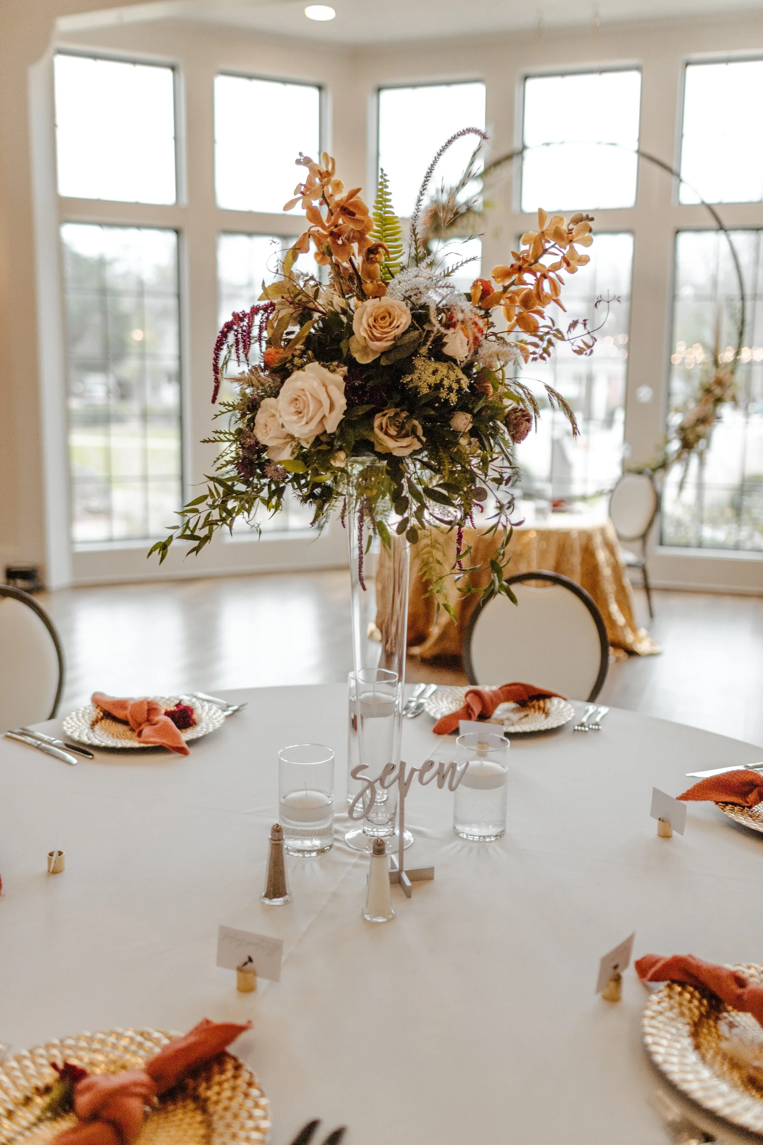 A round banquet table decorated with a tall floral centerpiece, place settings, and glasses of water in a bright room with large windows.
