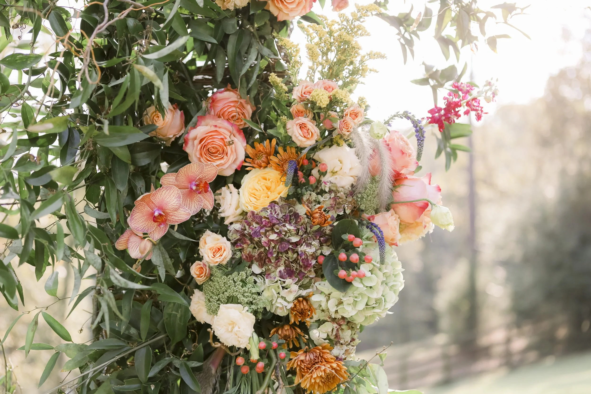 A floral arrangement of pink, orange, yellow, white, and purple flowers with green leaves, set outdoors with sunlight in the background.