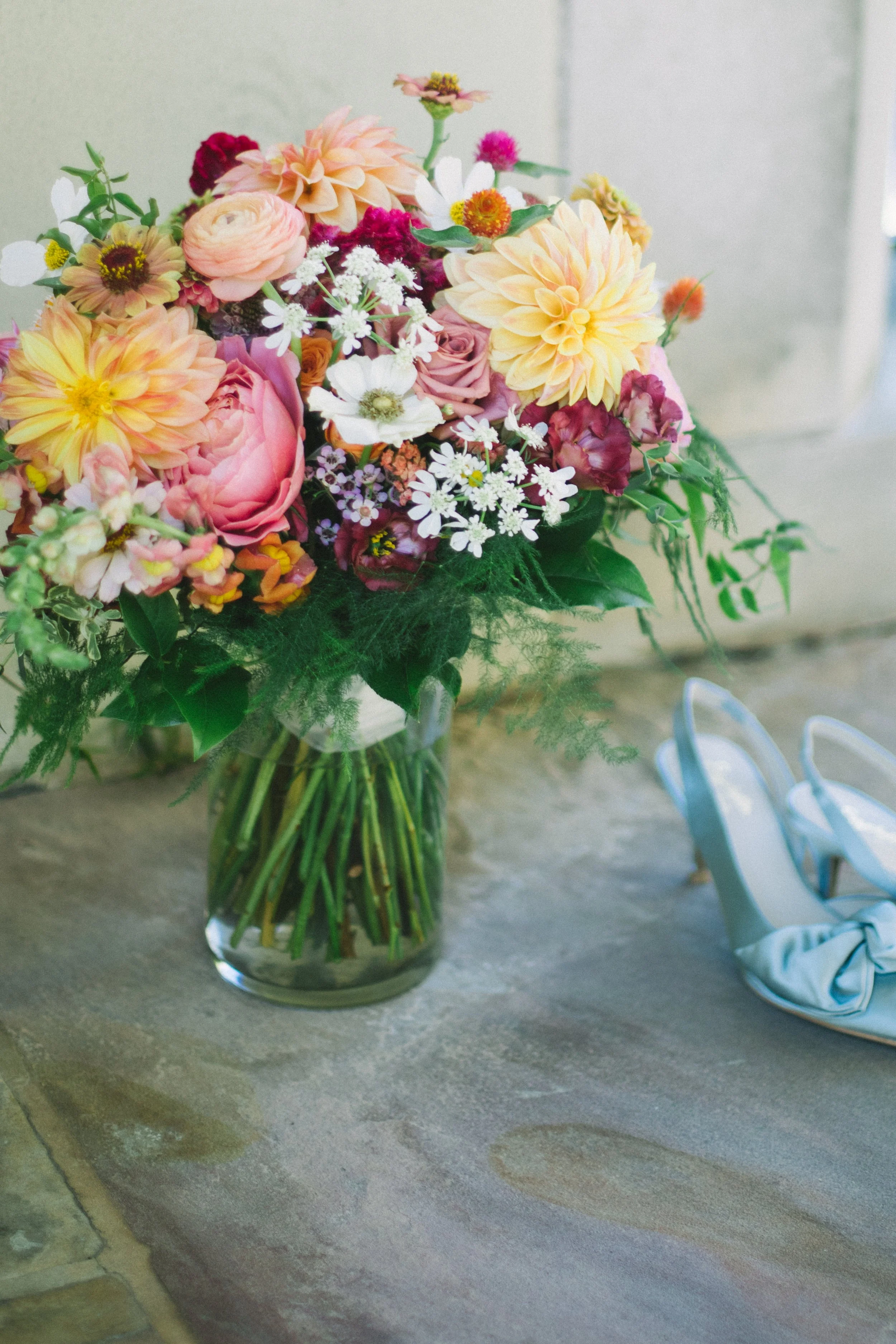 A colorful bouquet of assorted fresh flowers in a clear glass vase on a stone table, with a pair of light blue high-heeled shoes nearby.