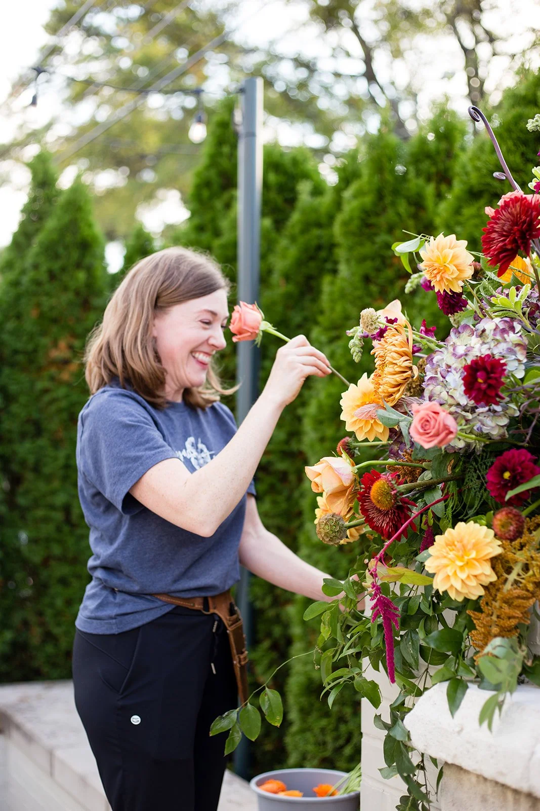 A woman is smiling while placing a pink rose in a large colorful flower arrangement outdoors.