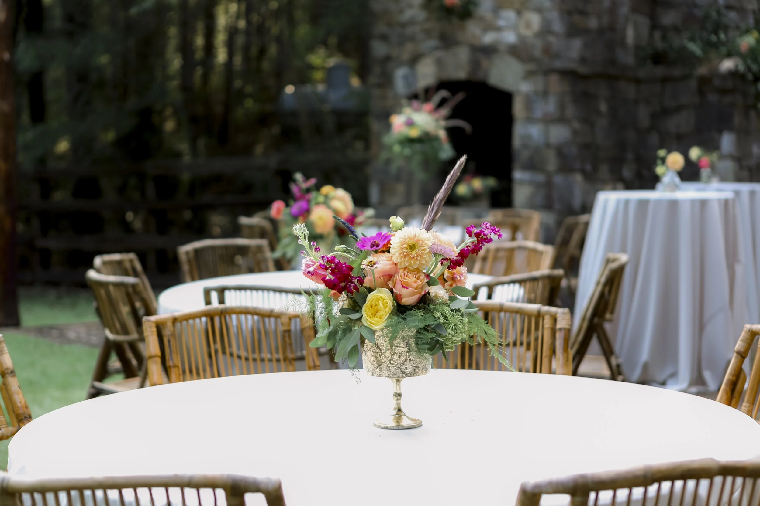 A floral centerpiece with pink, yellow, and purple flowers on a white tablecloth at an outdoor event, with wooden chairs and more floral arrangements in the background.
