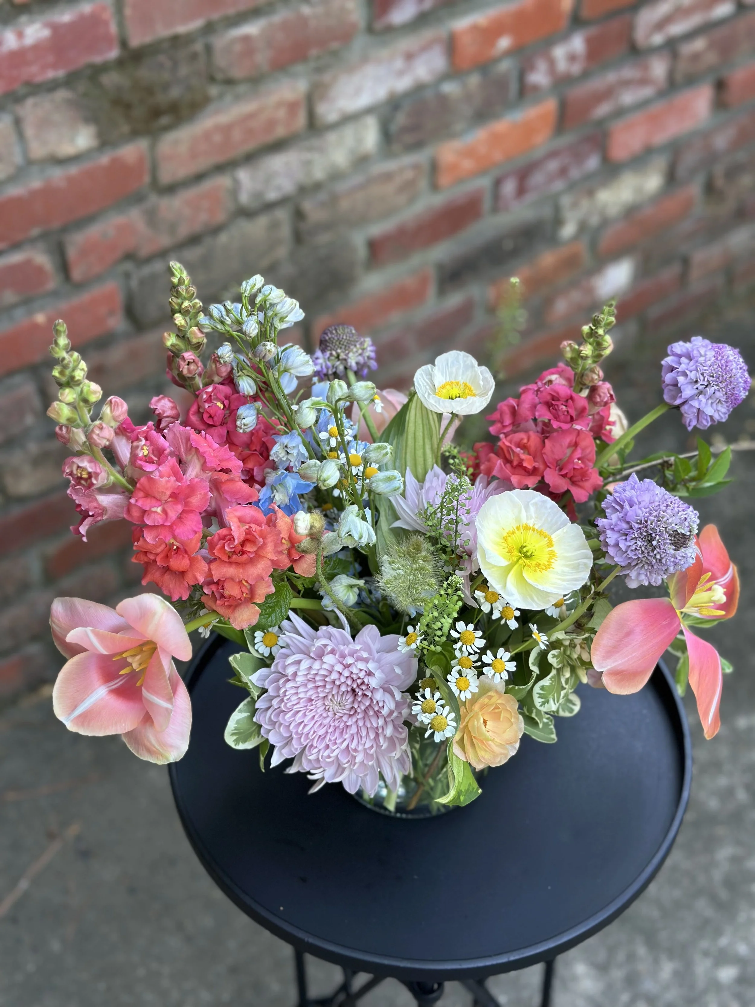Colorful bouquet of mixed flowers, including daisies, poppies, snapdragons, and other pastel-colored blossoms, on a black round table against a brick wall background.