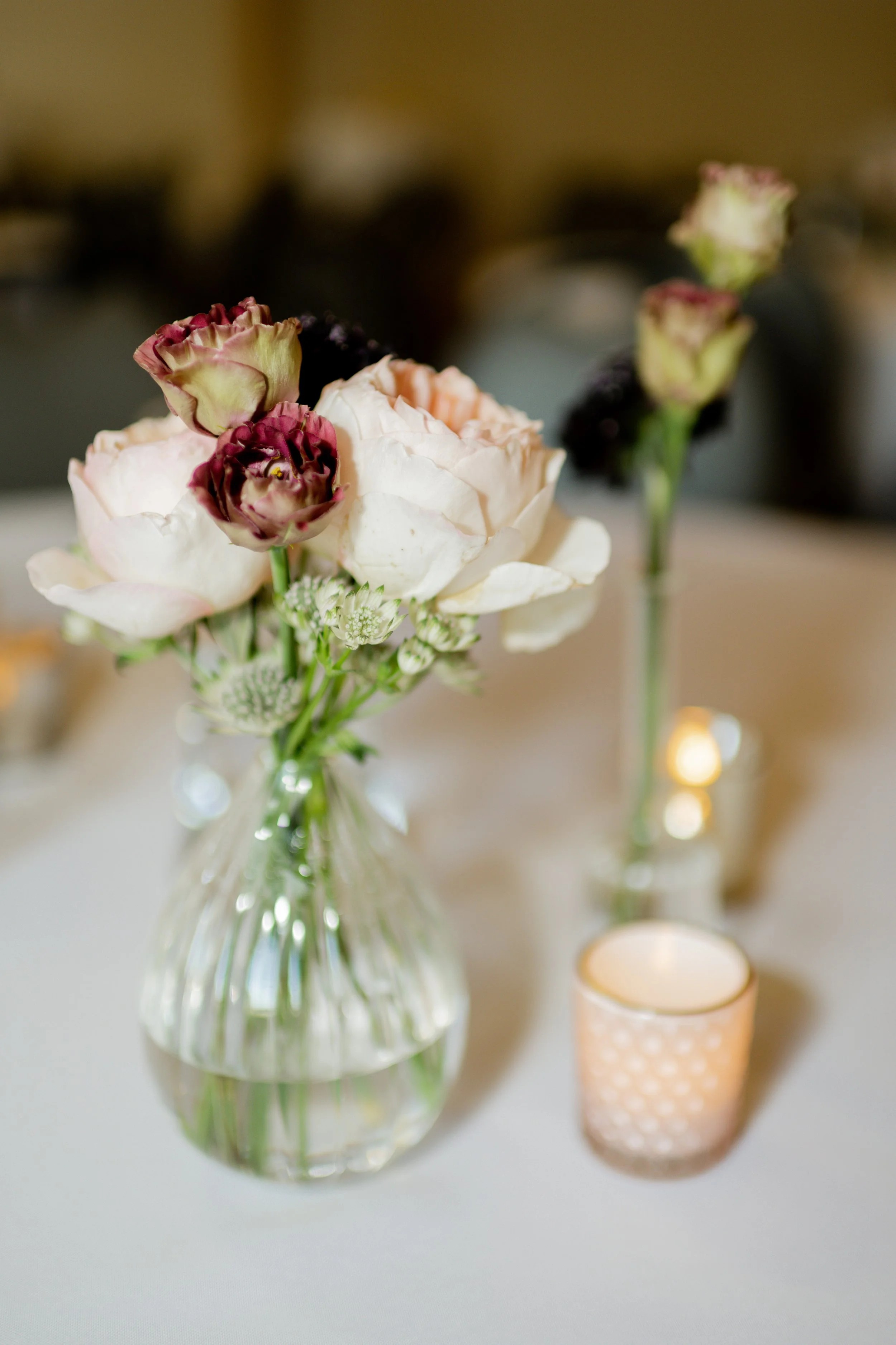 A floral arrangement in a glass vase with pink and white flowers, next to a small candle holder with a lit candle, on a white tablecloth.