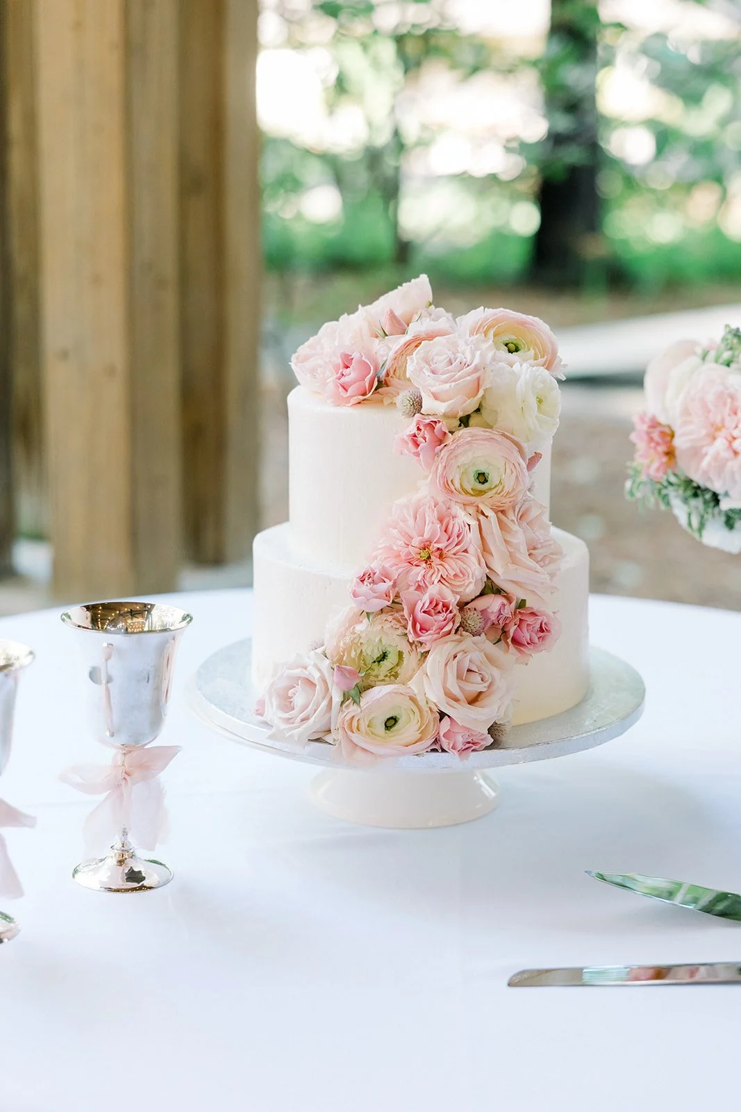 A two-tiered white wedding cake decorated with cascading pink and white flowers, placed on a table with silver cups and a cake knife in an outdoor setting.