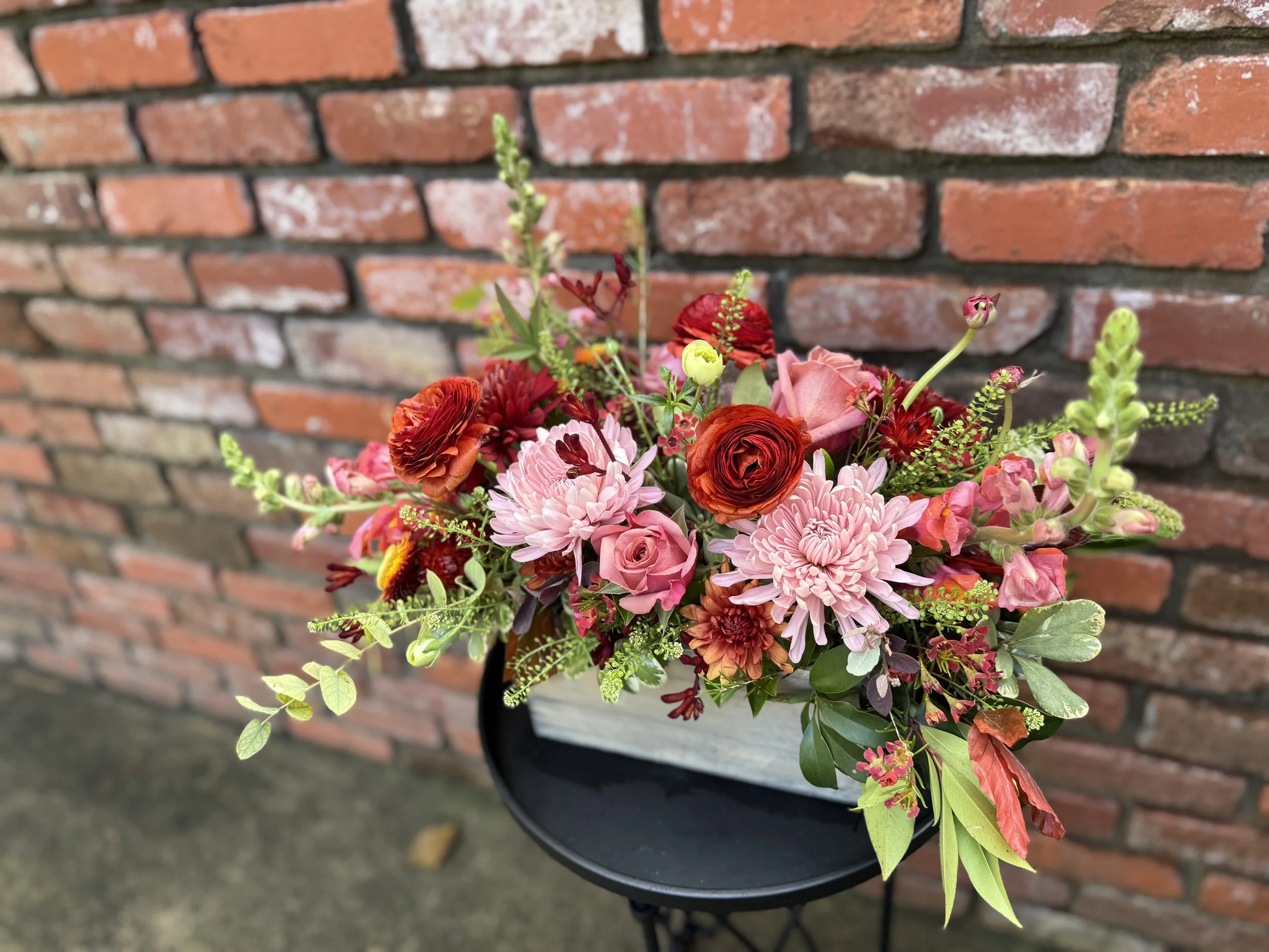 A colorful bouquet of pink, red, and orange flowers arranged in a black container on a black stand, set against a brick wall background.
