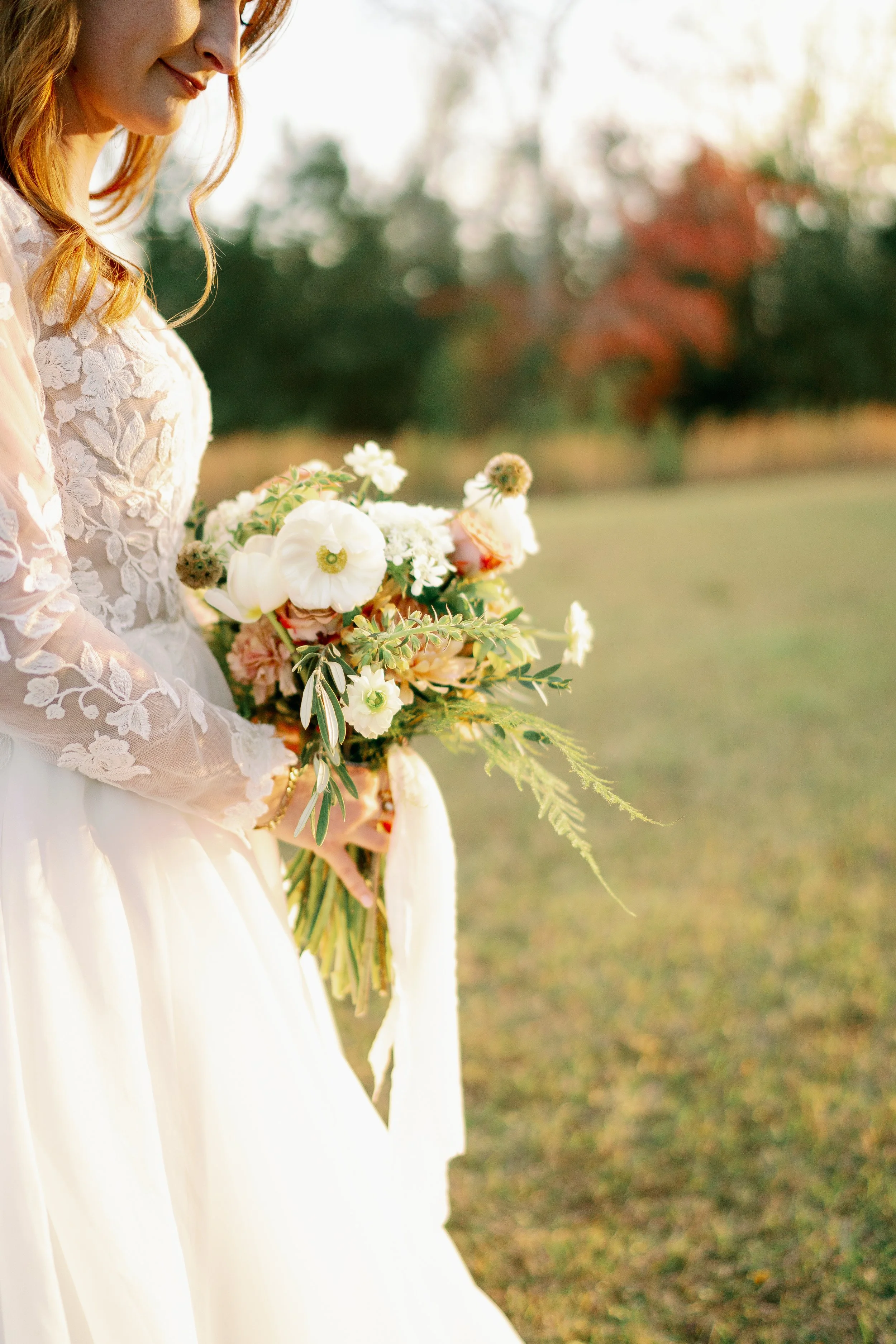 A woman wearing a white lace dress holding a bouquet of white and blush pink flowers outdoors in a grassy area with trees in the background during sunset.