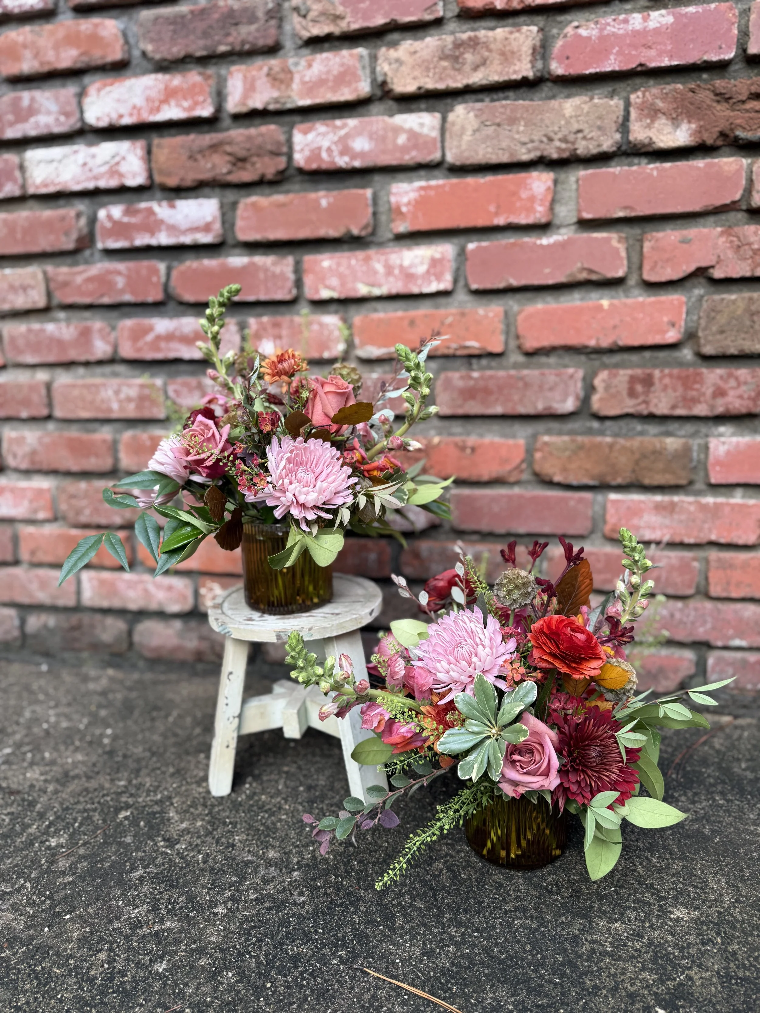 Two floral arrangements in amber glass vases on a small white wooden stool and on the ground, set against a brick wall background.
