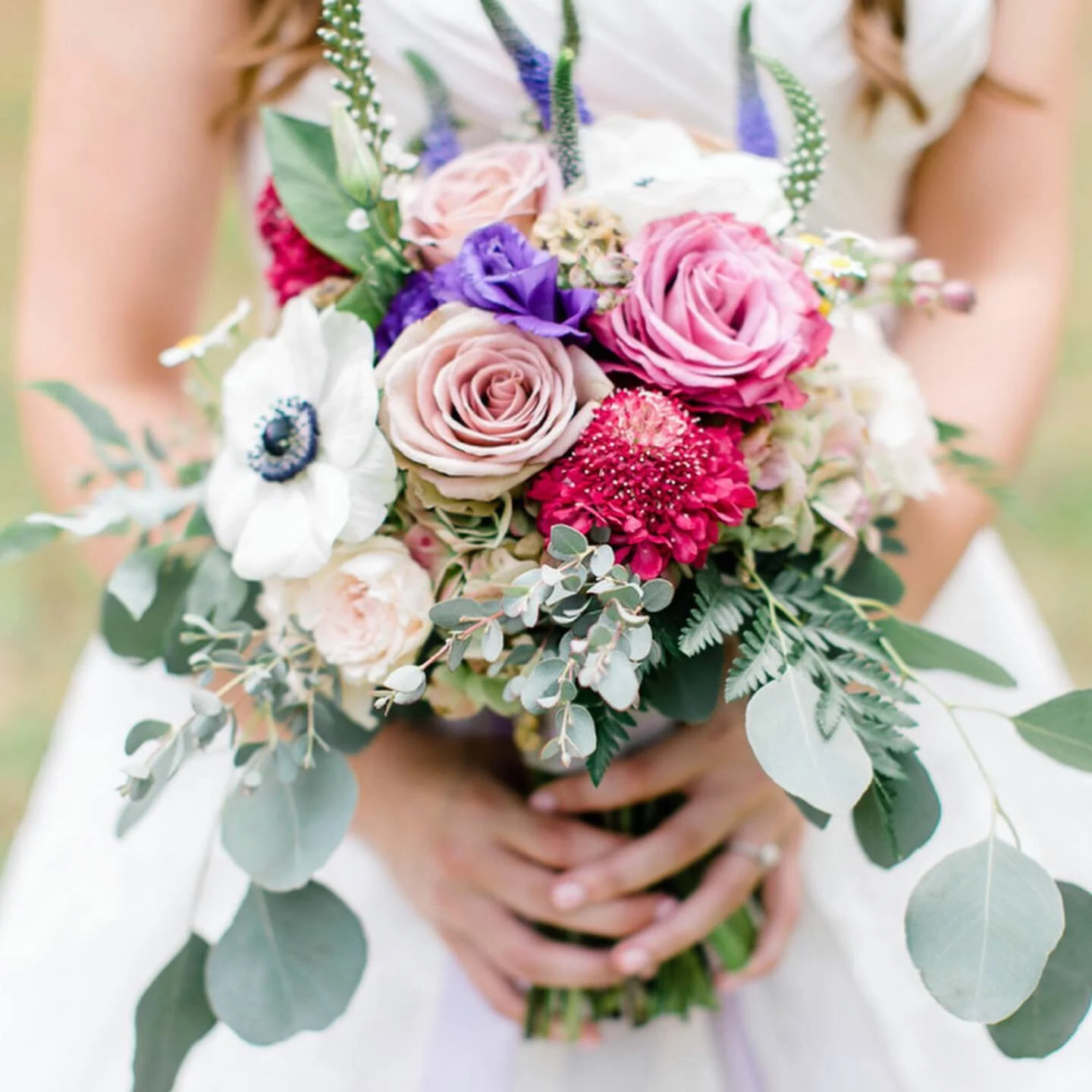 A person holding a bouquet of pink, purple, white, and red flowers with green foliage.