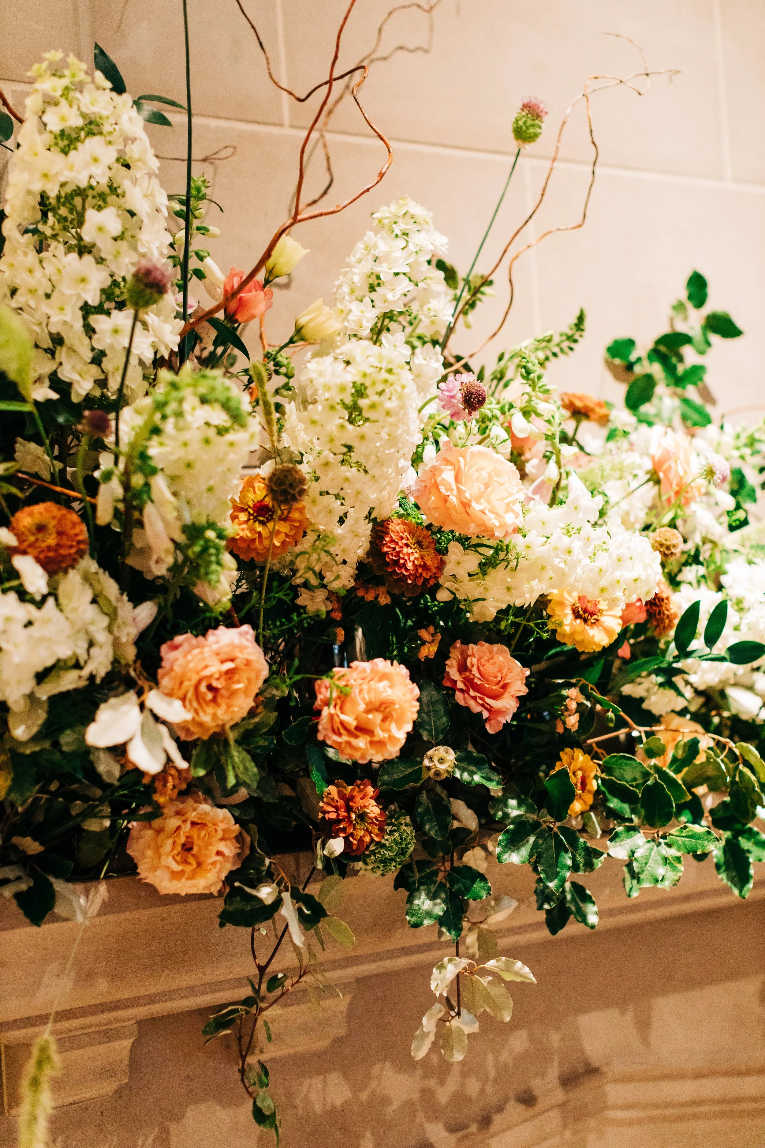 A vibrant flower arrangement featuring white hydrangeas, peach roses, orange daisies, and greenery, set against a neutral background.
