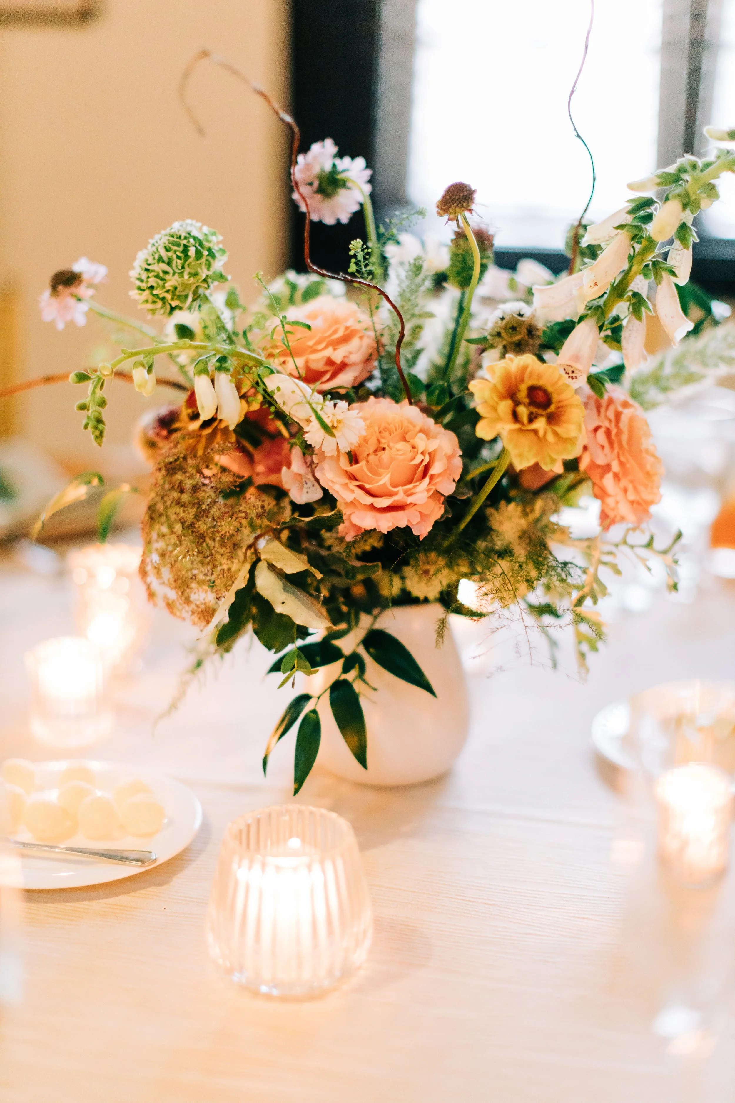 A floral arrangement with pink, peach, and cream-colored flowers in a white vase on a table with candles and a plate of cookies.