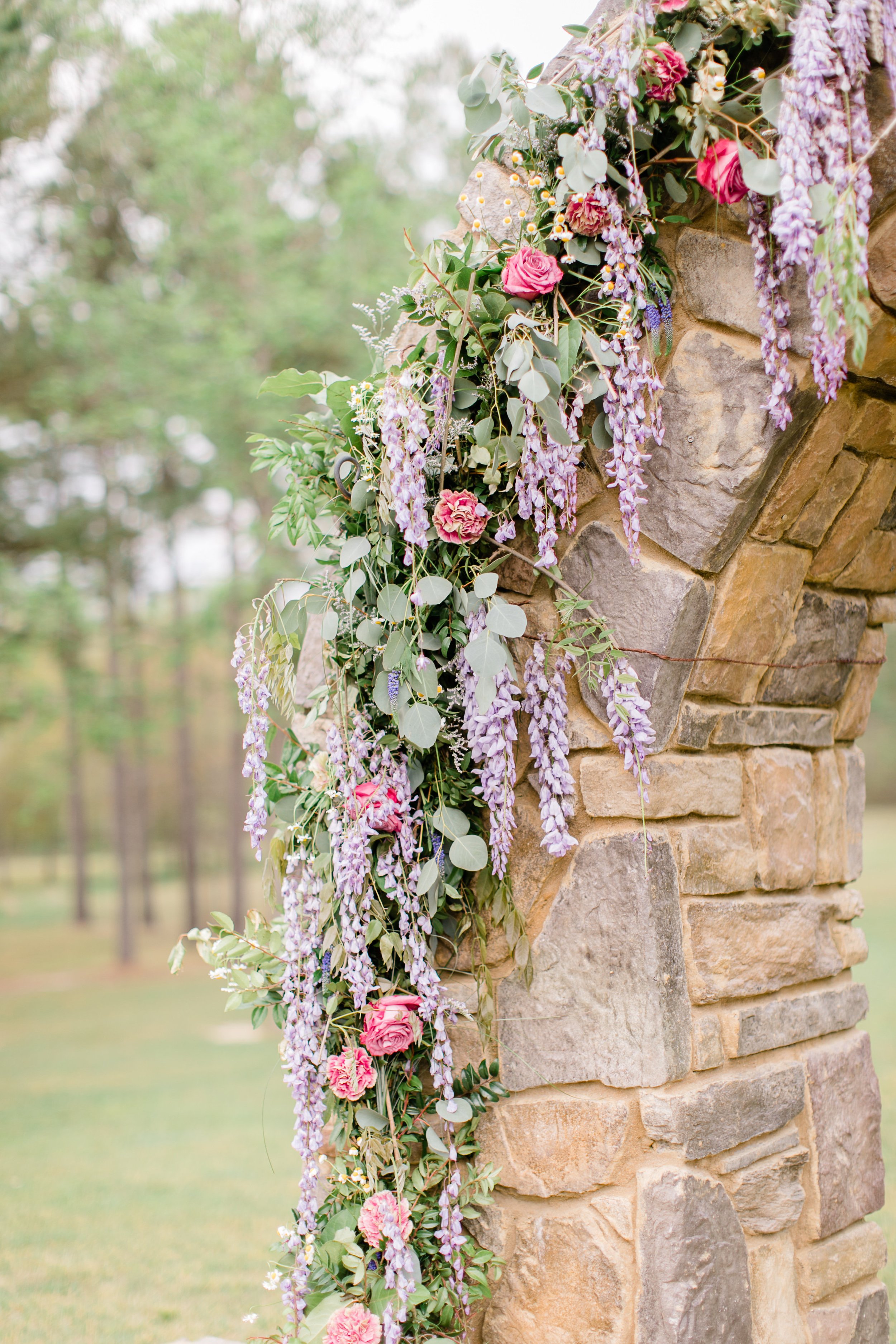 Decorative stone archway adorned with pink roses, purple wisteria, and green foliage outdoors.