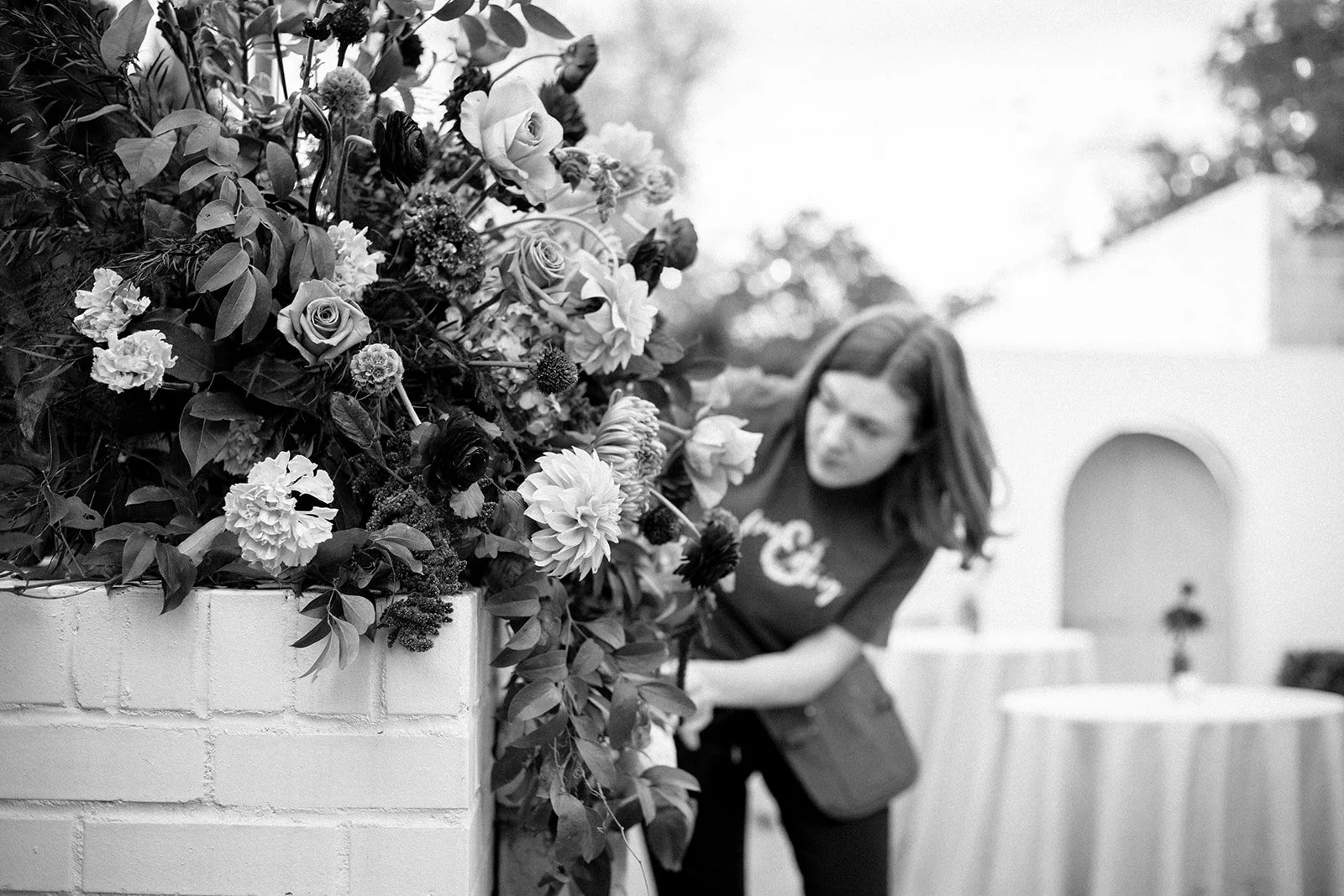 A woman arranging a large bouquet of mixed flowers next to a white brick wall outdoors.