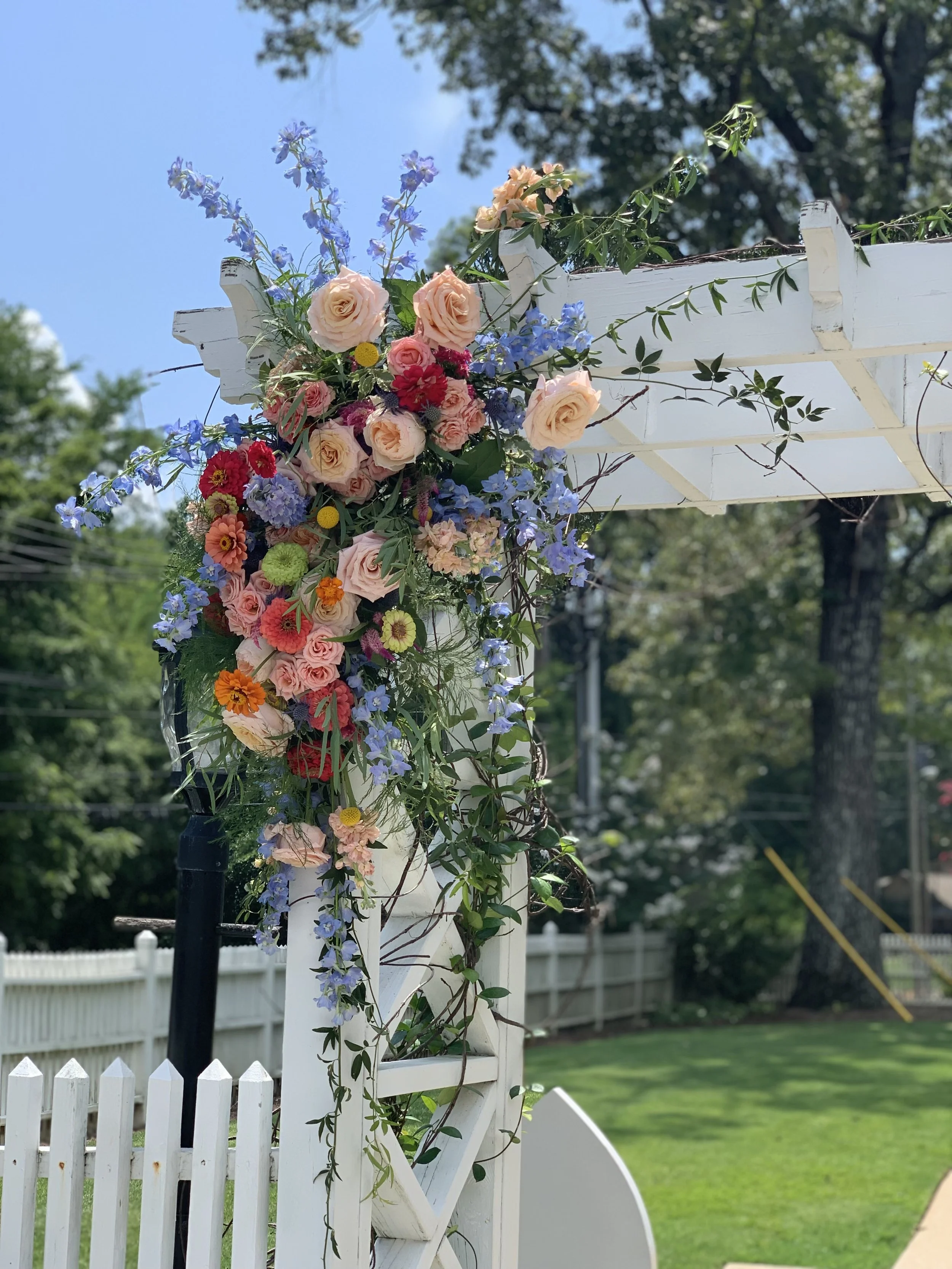 Wedding arch adorned with pink, purple, red, and orange flowers, including roses and delphiniums, outdoors on a sunny day.