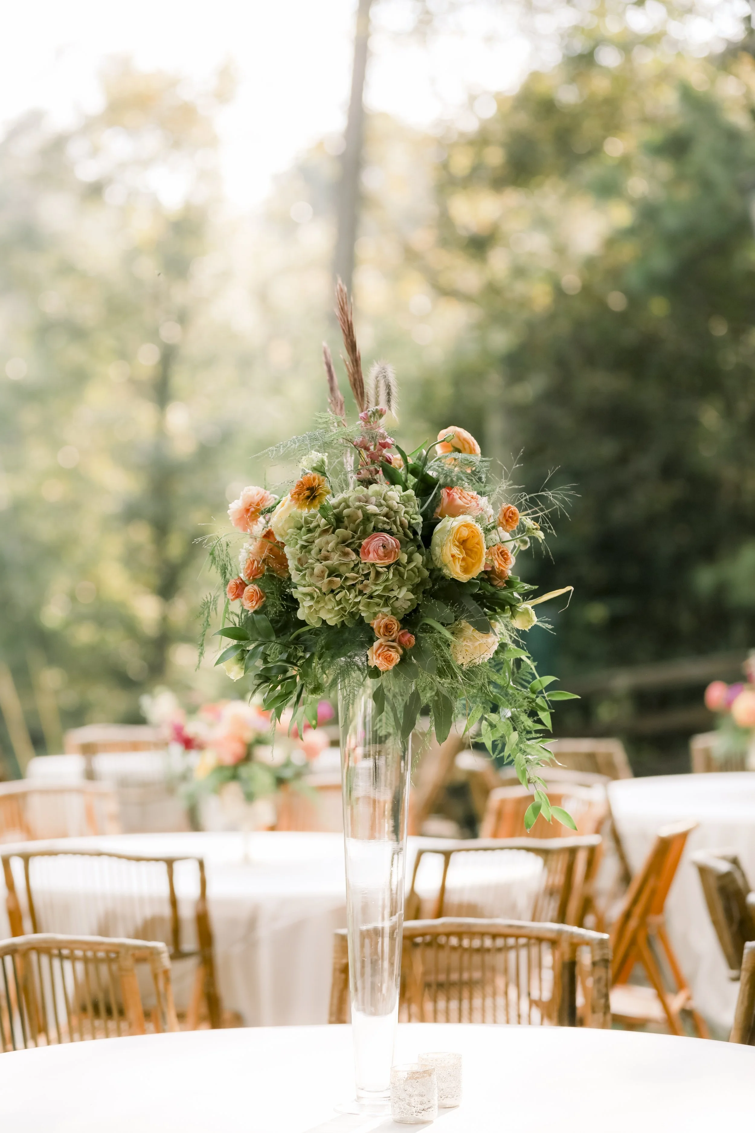 Tall floral centerpiece with peach and yellow roses, green hydrangeas, and greenery on a table at an outdoor event.