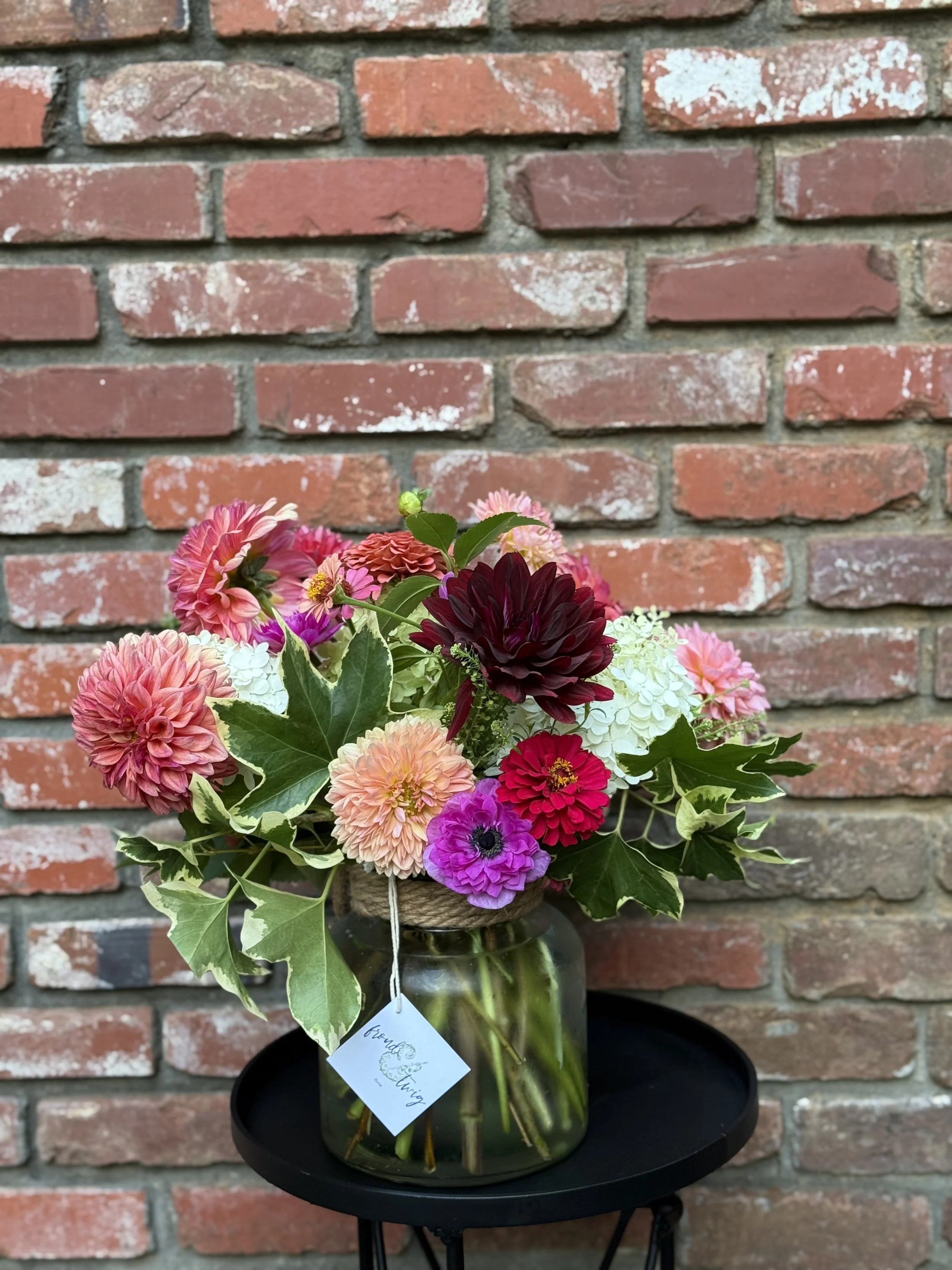A colorful bouquet of flowers including dahlias, anemones, and assorted greenery in a glass jar on a black round table against a brick wall.