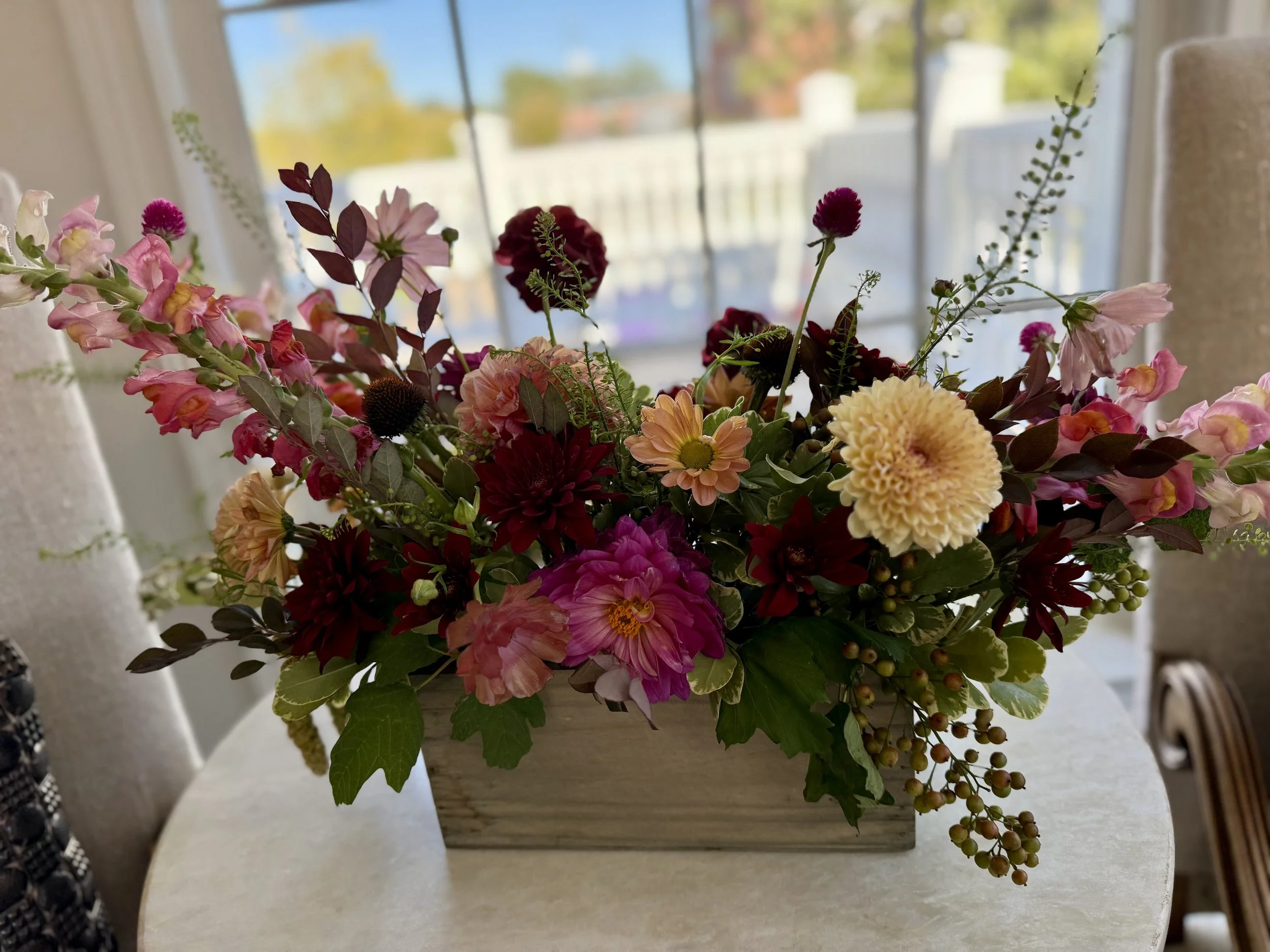 Arrangement of pink, red, orange, and yellow flowers in a wooden box on a round table, with a window and blurred outdoor scenery in the background.