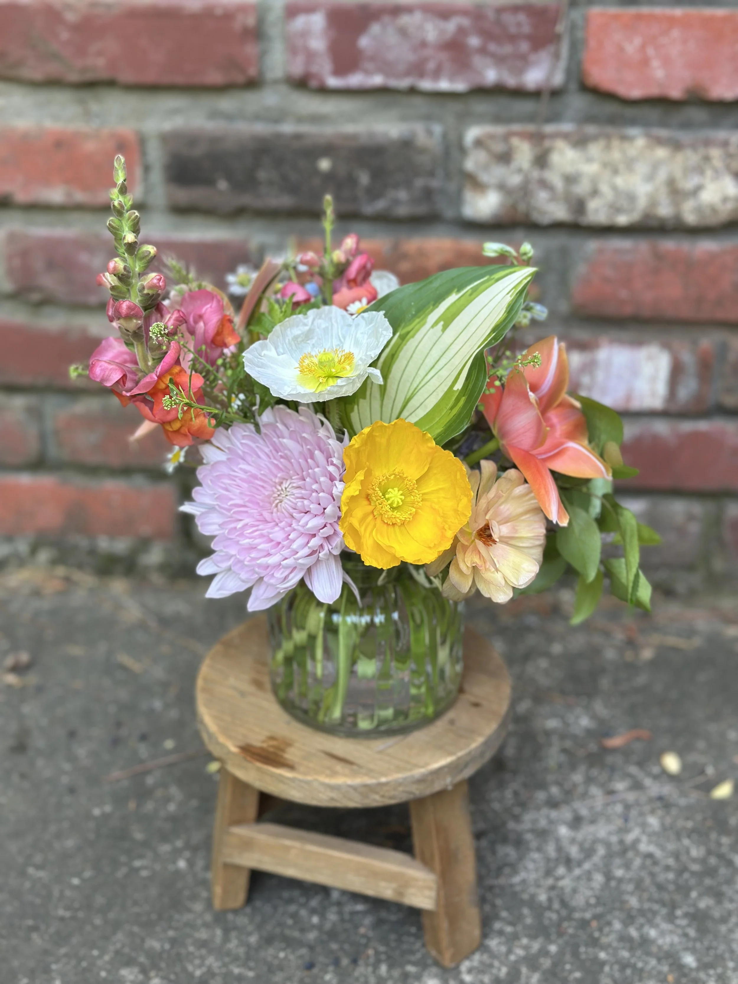 Colorful flower bouquet in a glass vase on a small wooden stool against a brick wall background.
