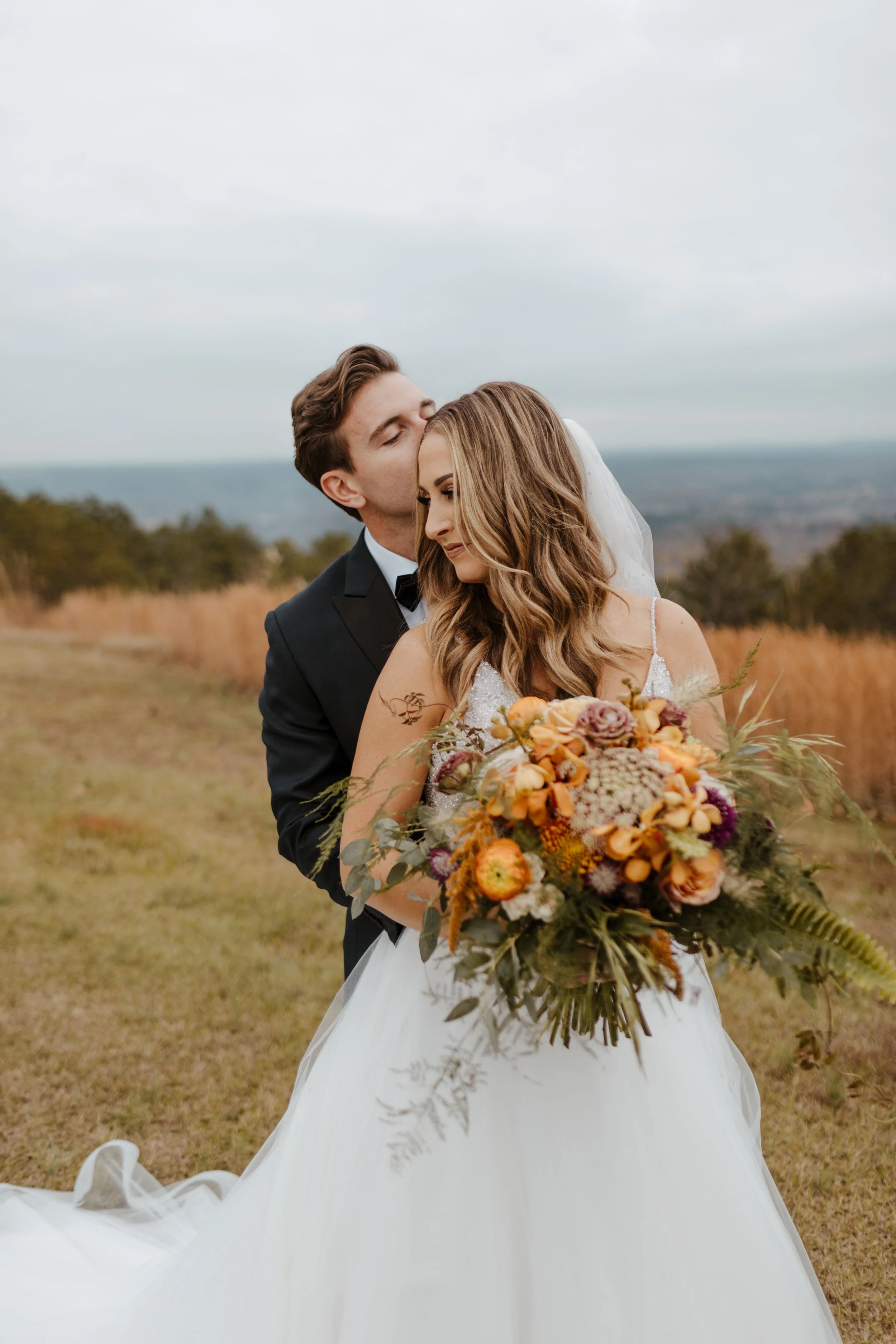 A bride and groom in an outdoor field, with the groom kissing the bride's forehead while holding a bouquet of flowers.