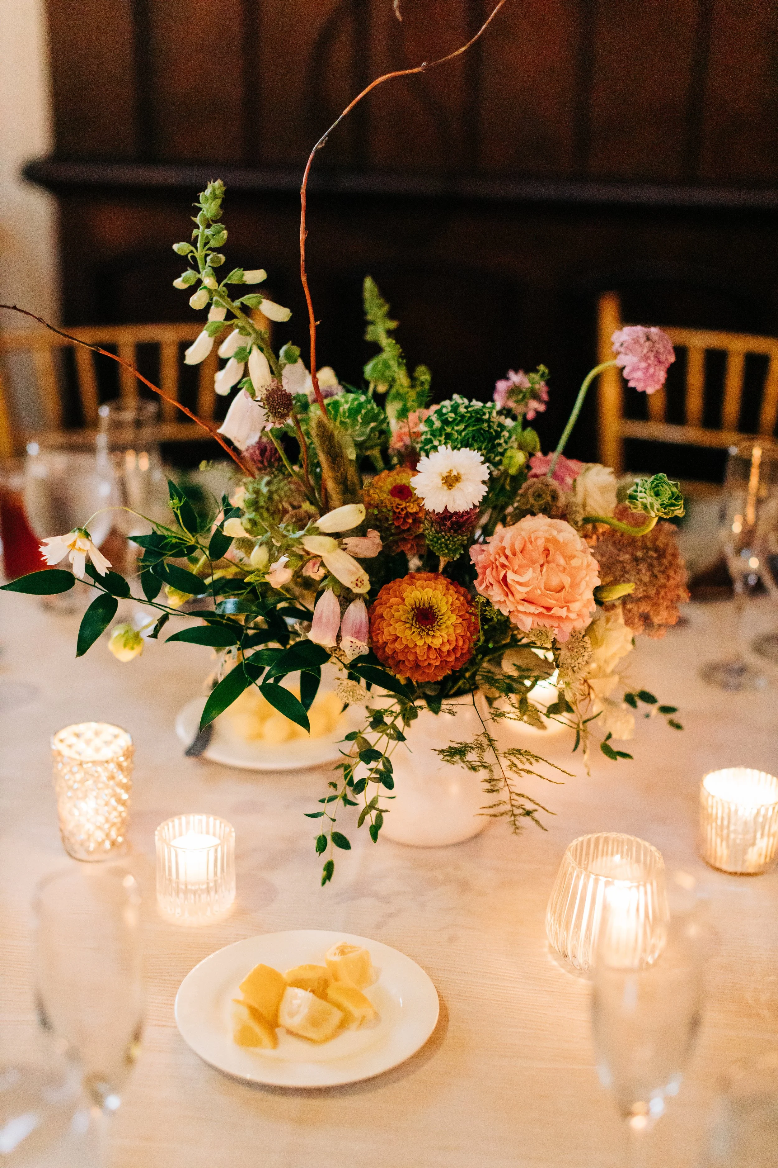 A floral centerpiece on a dining table with candles and cheese, set in a warmly lit room.