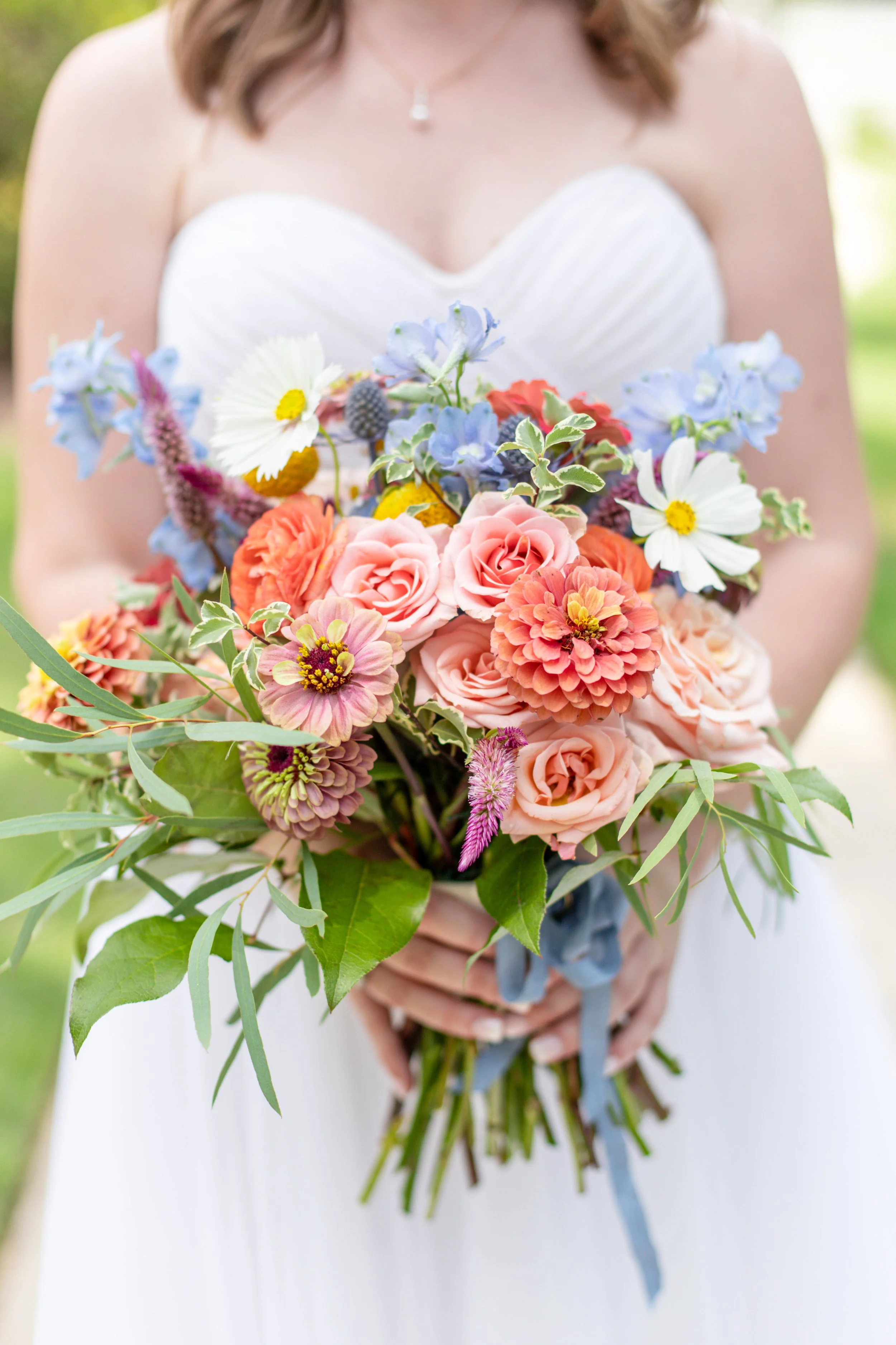 A woman in a white dress holding a colorful bouquet of various flowers including roses, daisies, and other blooms, with green leaves and a blue ribbon.
