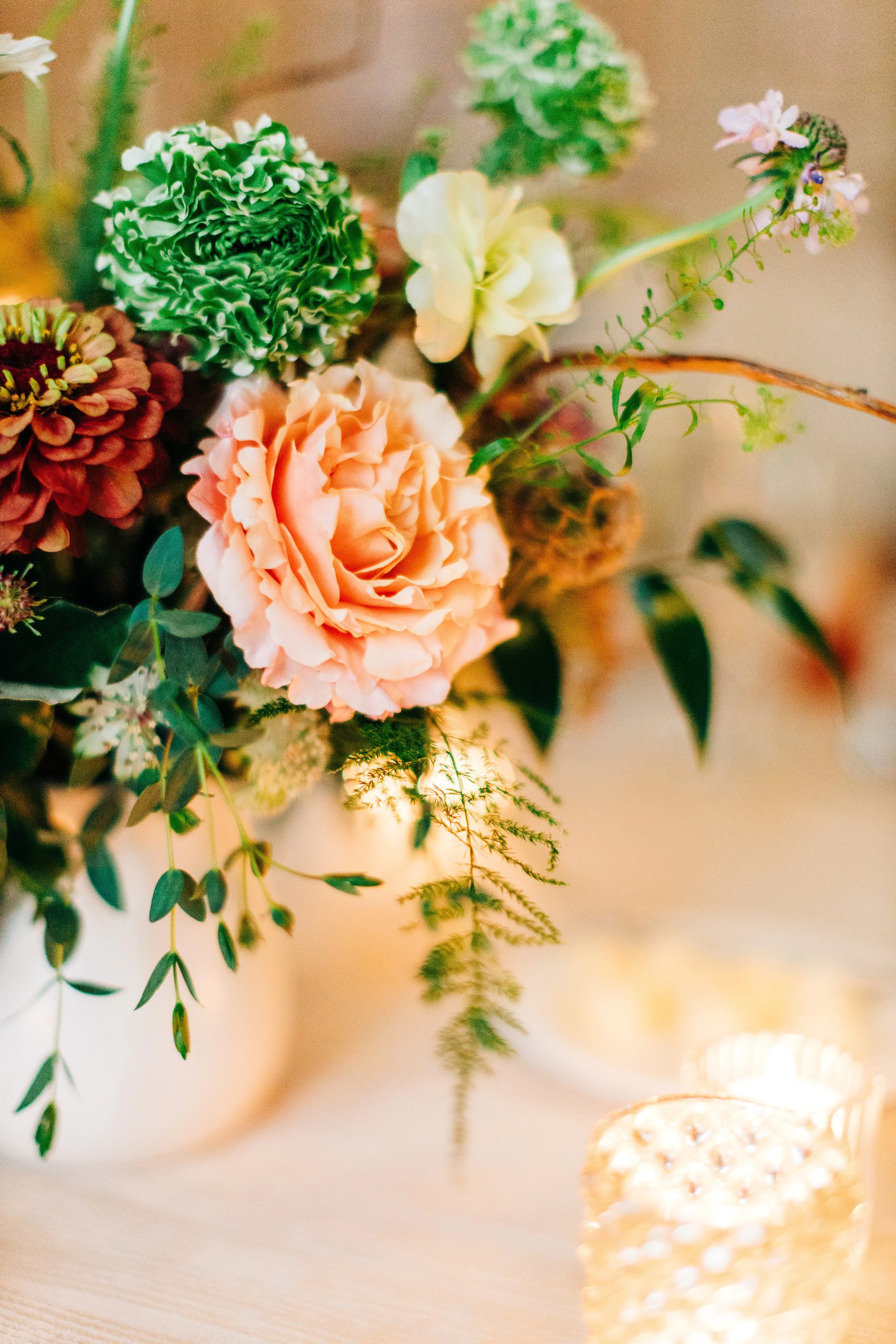 A floral arrangement with various flowers including a pink rose, green and white carnations, and small pink and white blossoms, with greenery in a white vase. Candles are lit nearby.