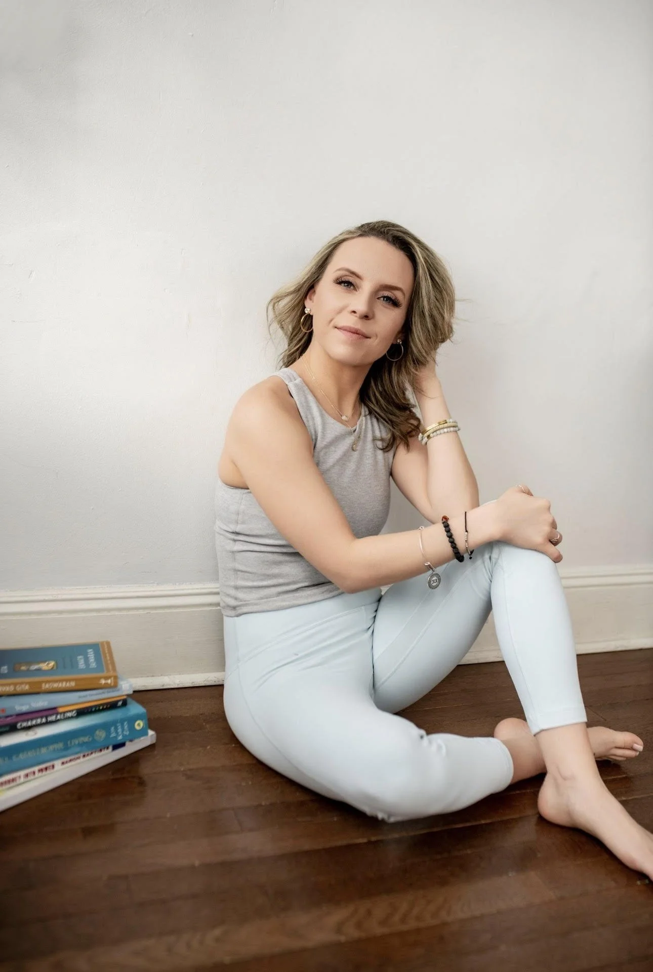 Picture of owner of Lauren Daly Clark Yoga & Wellness, LLC.  White female with blonde hair and blue eyes wearing light blue yoga pants and a gray top seated next to a small pile of books.