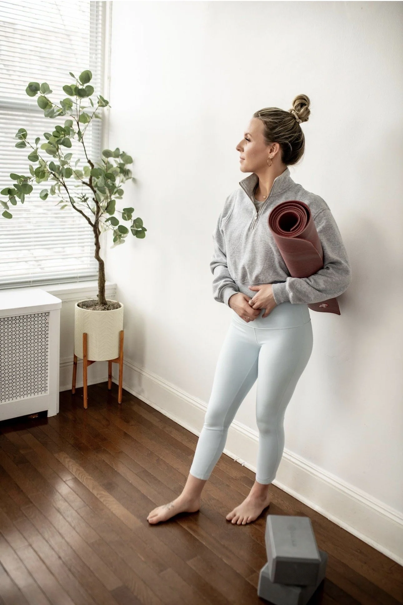 Picture of Lauren, owner of Lauren Daly Clark Yoga & Wellness, LLC.  Lauren is standing against a wall with a yoga mat under her arm and yoga blocks by her feet.  She is wearing light blue yoga pants and gray cropped sweatshirt.