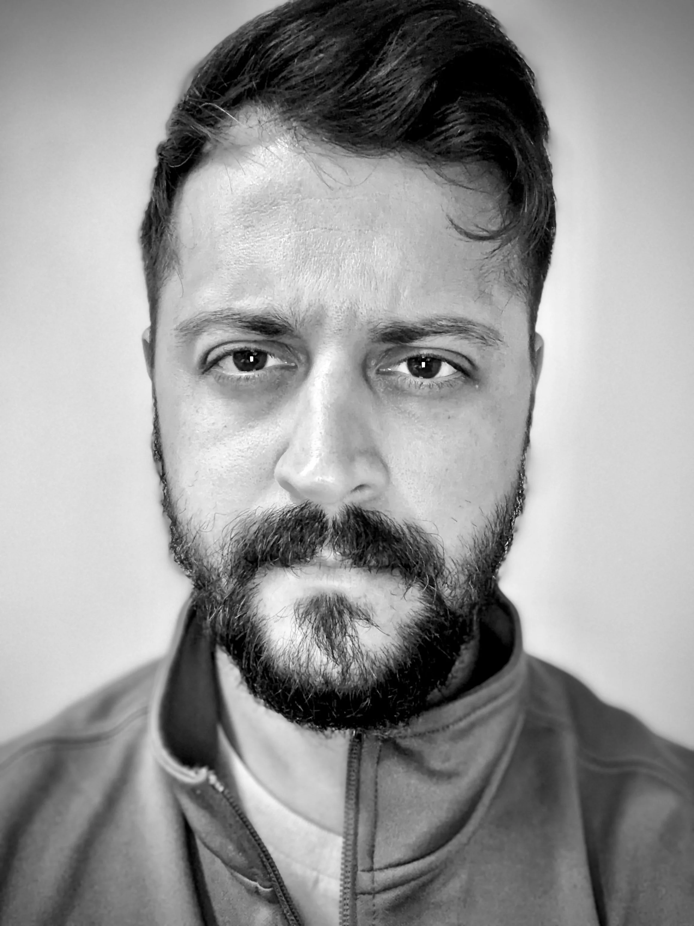 Black and white portrait of a man with dark hair, a beard, and mustache, wearing a zip-up jacket, looking slightly to the side.
