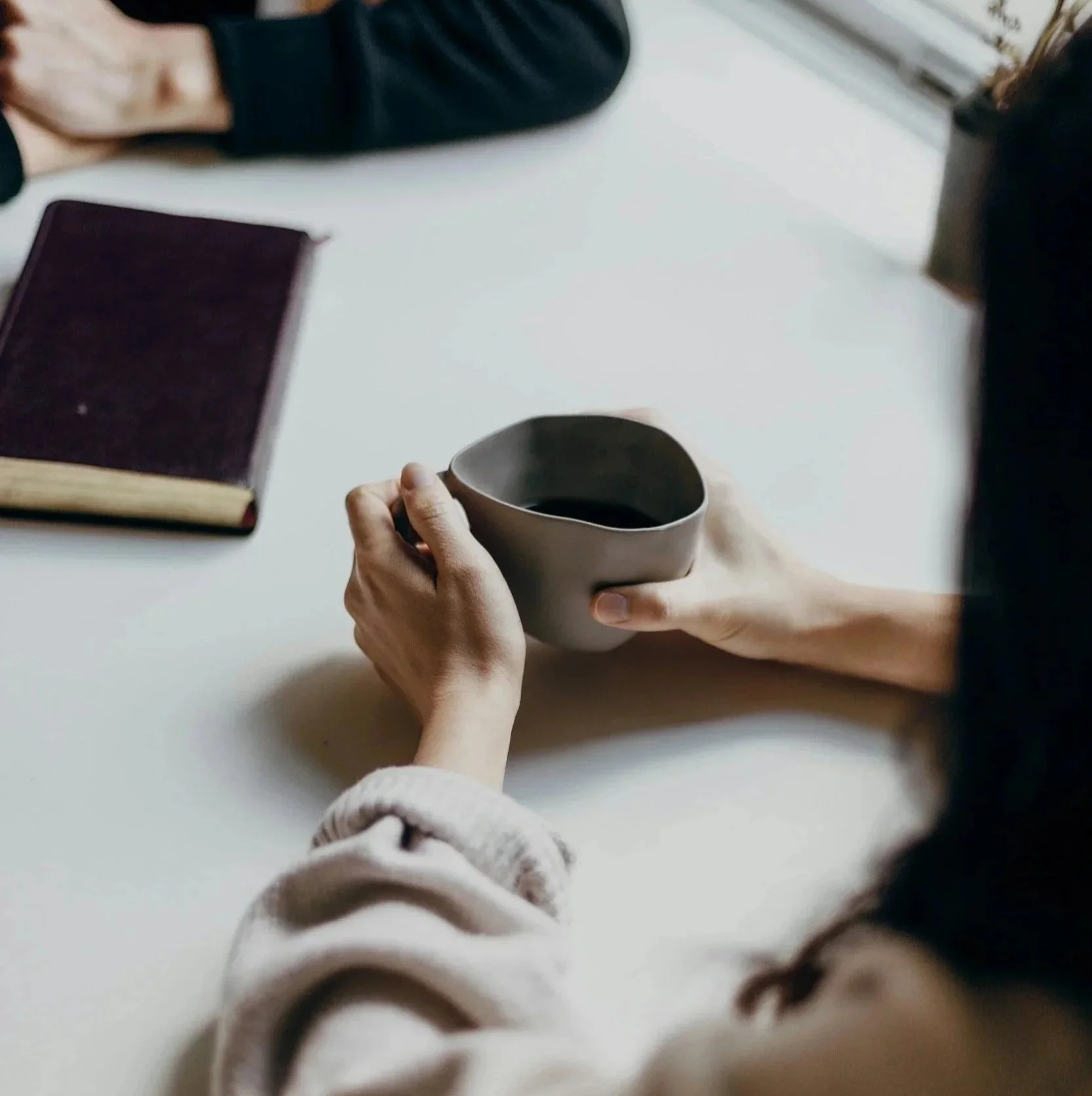 Person hält eine dunkel gefärbte Tasse mit schwarzem Kaffee. Auf dem weißen Tisch liegt ein Buch mit dunkelrotem Umschlag. Es ist eine ruhige Szene, wahrscheinlich im Inneren eines Hauses.