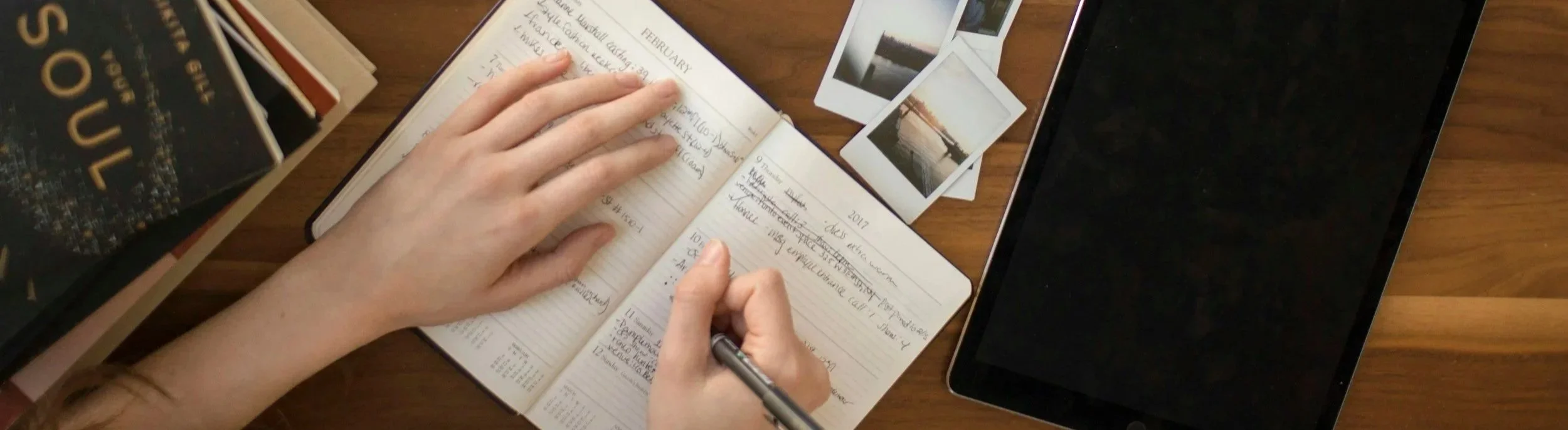 Eine Person schreibt in einem schwarz umrahmten Tagebuch, umgeben von Fotos, Büchern und einem Tablet auf einem Holzschreibtisch.