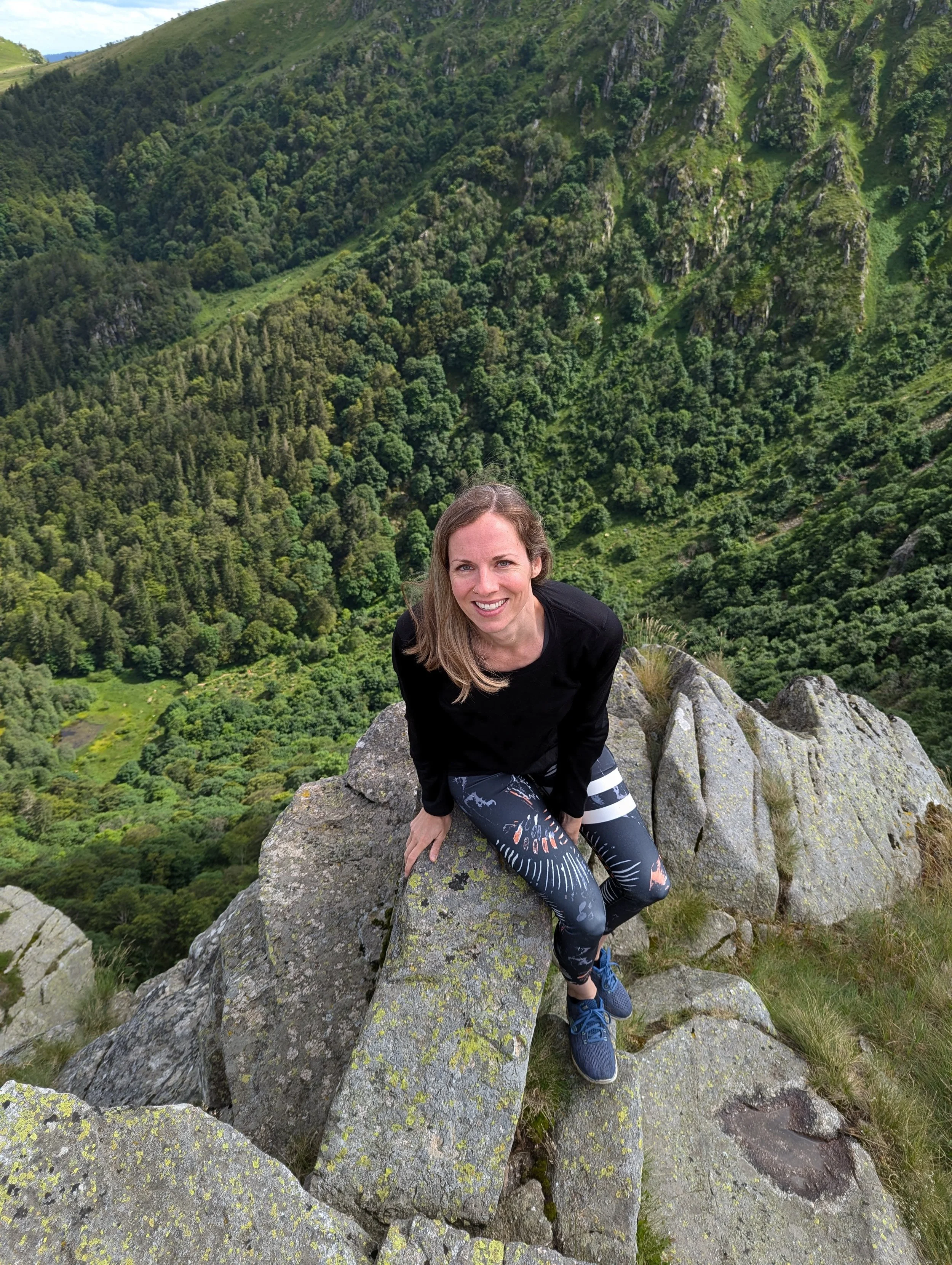 Frau sitzt auf einem Felsvorsprung mit Blick auf einen grünen, bewaldeten Hügel.
