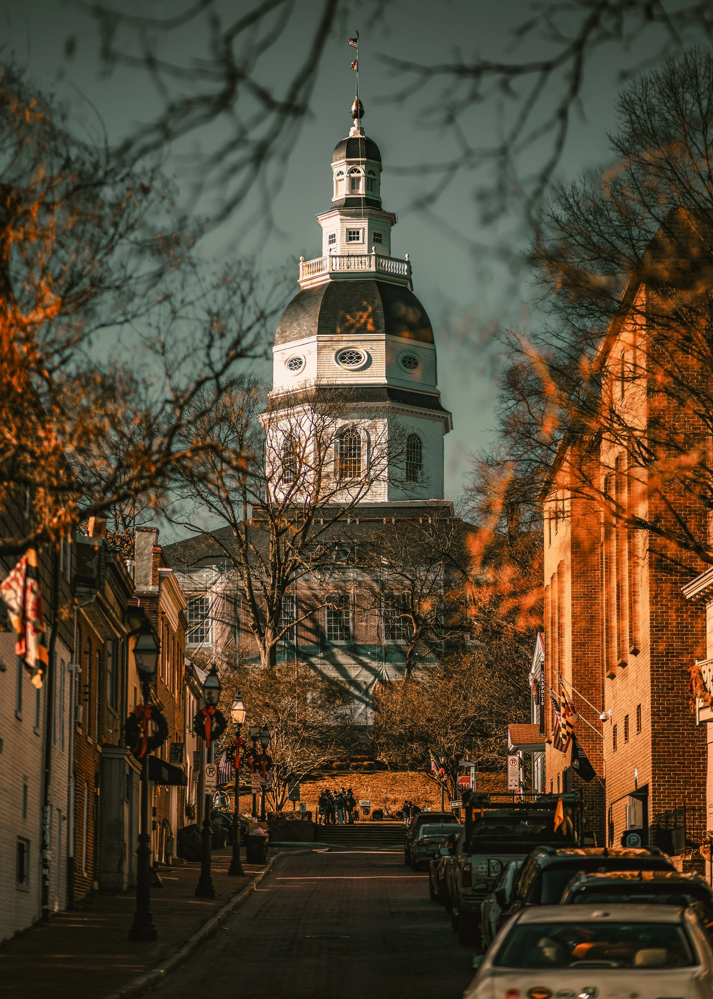 A city street with parked cars, storefronts on both sides, and a church steeple in the background, taken during early evening or late afternoon.